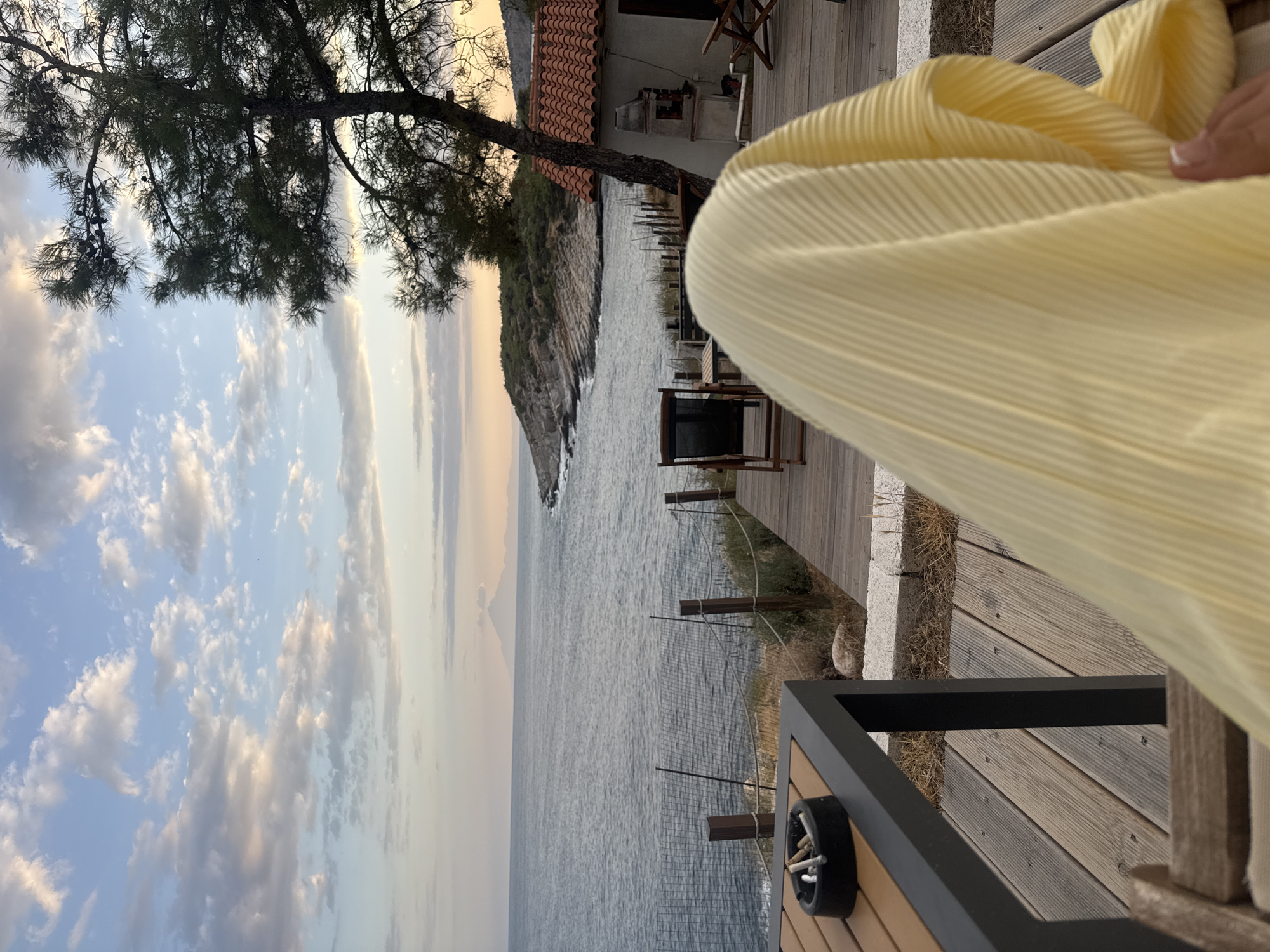 The image depicts a tranquil scene at dusk on a wooden deck overlooking the Aegean Sea in Greece. In the foreground, a close-up shot focuses on a yellow pleated fabric draped over a person's lap, with a hand reaching out towards it. To the left, a dark metal table holds an ashtray with several cigarette butts. Beyond the deck, the calm, grey sea stretches towards the horizon, where a faint, conical mountain is visible. The coastline to the right is rocky and covered in low-lying vegetation, with a small white building featuring a red-tiled roof. A tall pine tree stands to the right of the building, its branches silhouetted against the soft, pastel-colored sky filled with scattered clouds. The overall atmosphere is one of peaceful relaxation and natural beauty.