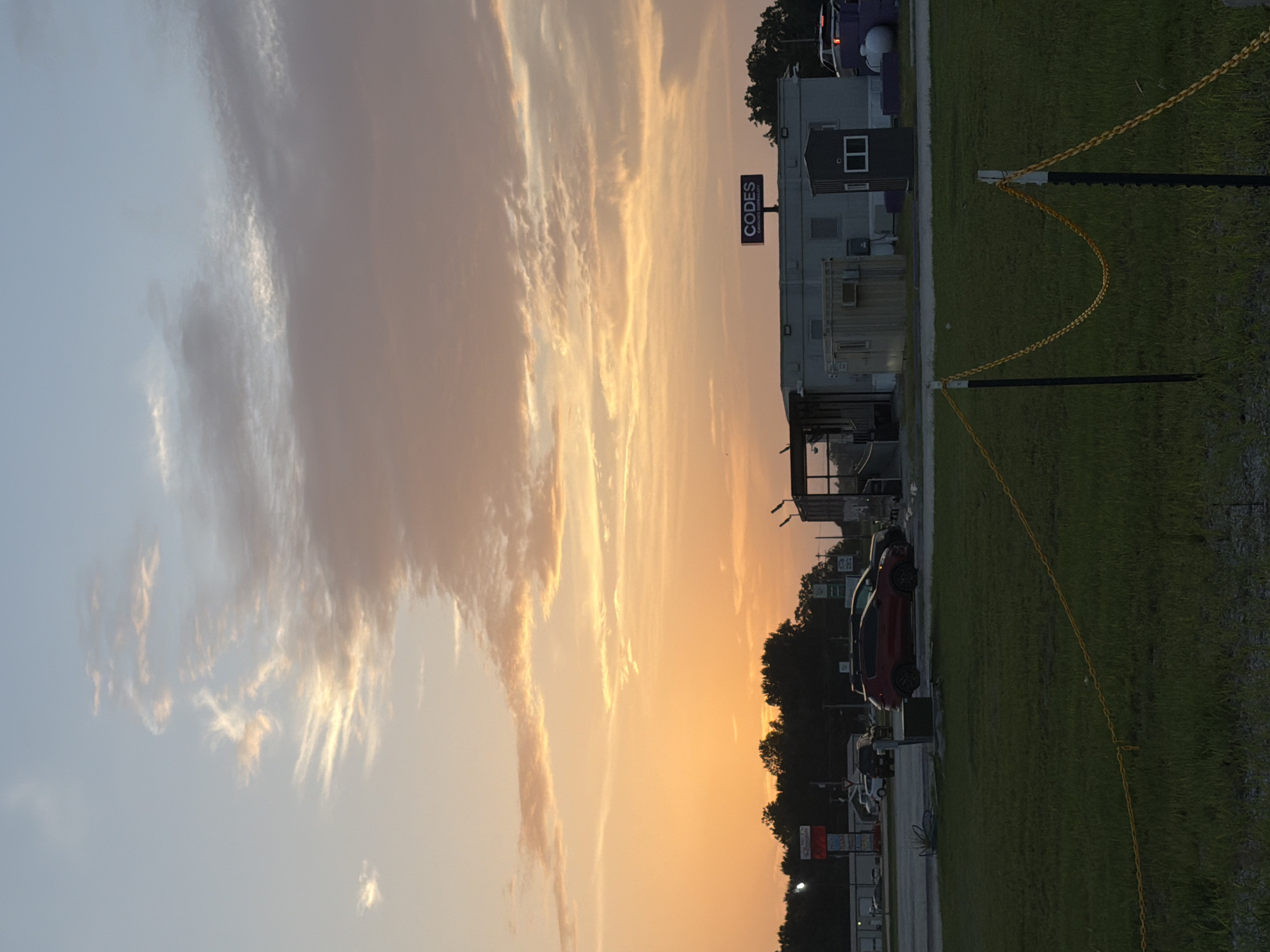 This image captures a sunset scene in Cardwell, United States. The sky is dominated by hues of orange, yellow, and pink, with scattered clouds. In the foreground, a grassy field is sectioned off by a yellow chain attached to black posts. Beyond the field, a parking lot is visible with several vehicles, including a red SUV and a black SUV. A long, grey mobile building complex stands in the middle ground, with a small guardhouse or booth to its right. A prominent sign with the word "CODES" in large purple letters and "CANNABIS DISPENSARY" in smaller white letters is mounted on top of the building. In the background, a line of trees separates the foreground from the sky. The overall impression is a commercial area at dusk.