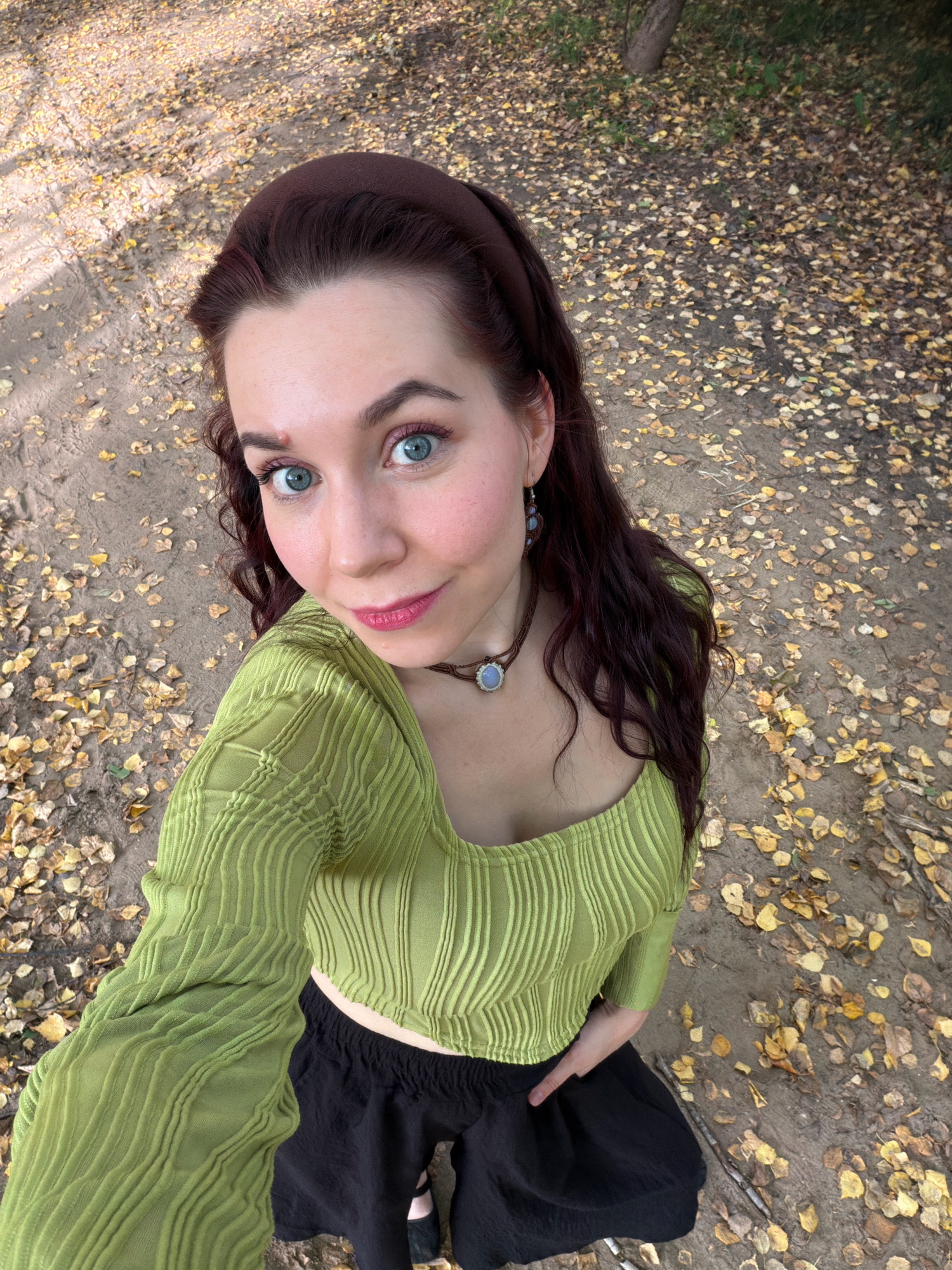 A young woman with long, dark, wavy hair and blue eyes is captured in a selfie from a high-angle perspective. She is wearing a lime green, textured long-sleeved crop top and black shorts. She has on a brown choker necklace with a blue pendant and matching earrings. The background is a dirt path covered in fallen yellow and brown leaves, suggesting an autumn setting. Sunlight filters through unseen trees, casting dappled shadows on the ground. The location is identified as Chaikovsky, Russia.