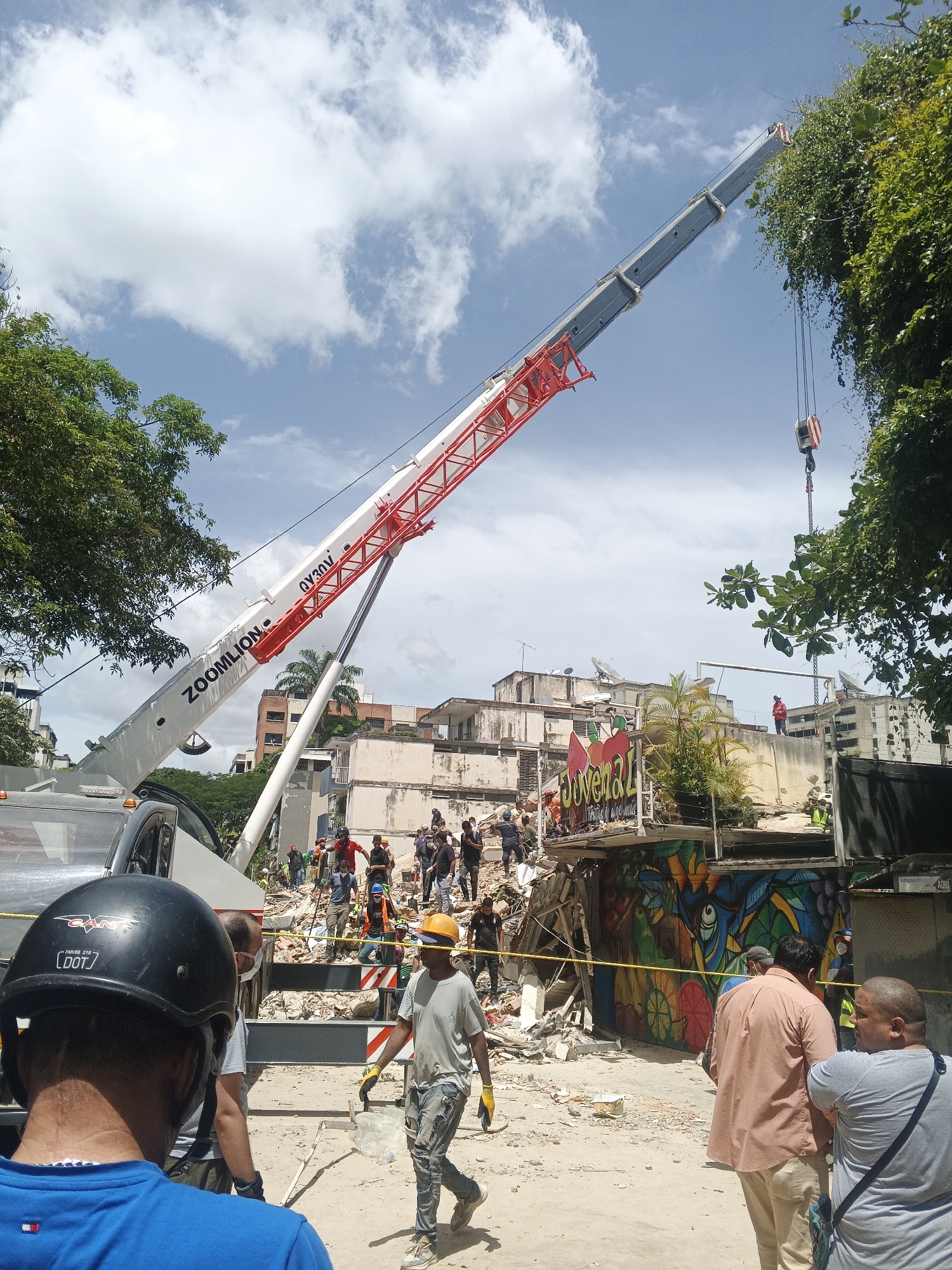 This media file depicts a scene of a partially collapsed building, likely a result of demolition or an accident, in Caracas, Venezuela. A large red and white crane, identified by the text "ZOONLION QY30V," dominates the upper portion of the image, its boom extending over the debris. Several individuals, some wearing hard hats and high-visibility vests, are present at the site, suggesting rescue or demolition operations. A truck is visible on the left, and a colorful mural adorns a surviving section of the building's wall, featuring an eye motif and fruit imagery. The sky is bright blue with scattered white clouds, indicating a daytime scene under fair weather conditions. The overall atmosphere appears to be one of ongoing work and assessment of the damaged structure.