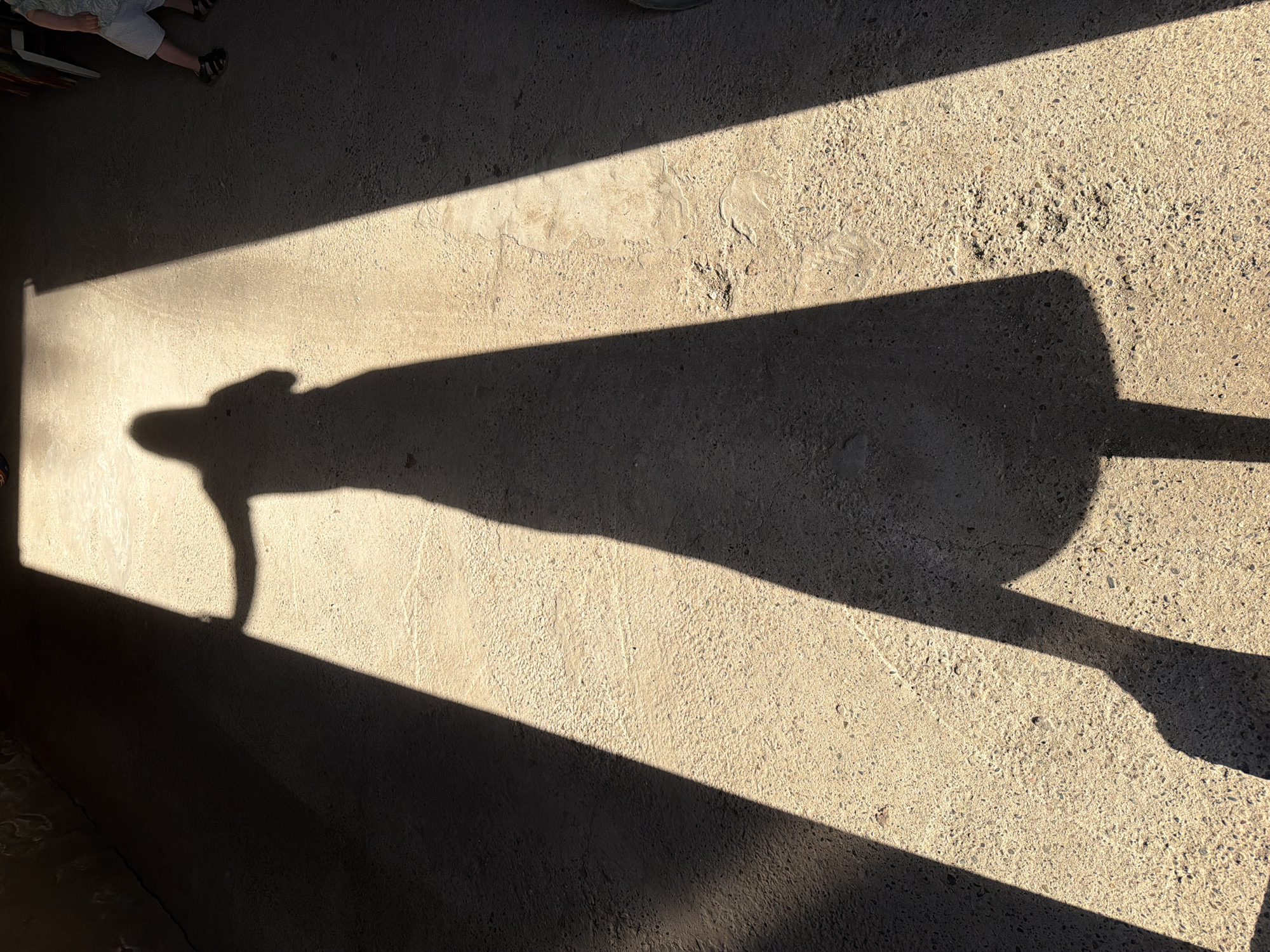 A shadow of a person is cast on a concrete surface divided by a beam of light. The shadow's head is on the left with legs extending to the right. The location is unknown and cannot be determined to be in Votkinsk, Russia.
