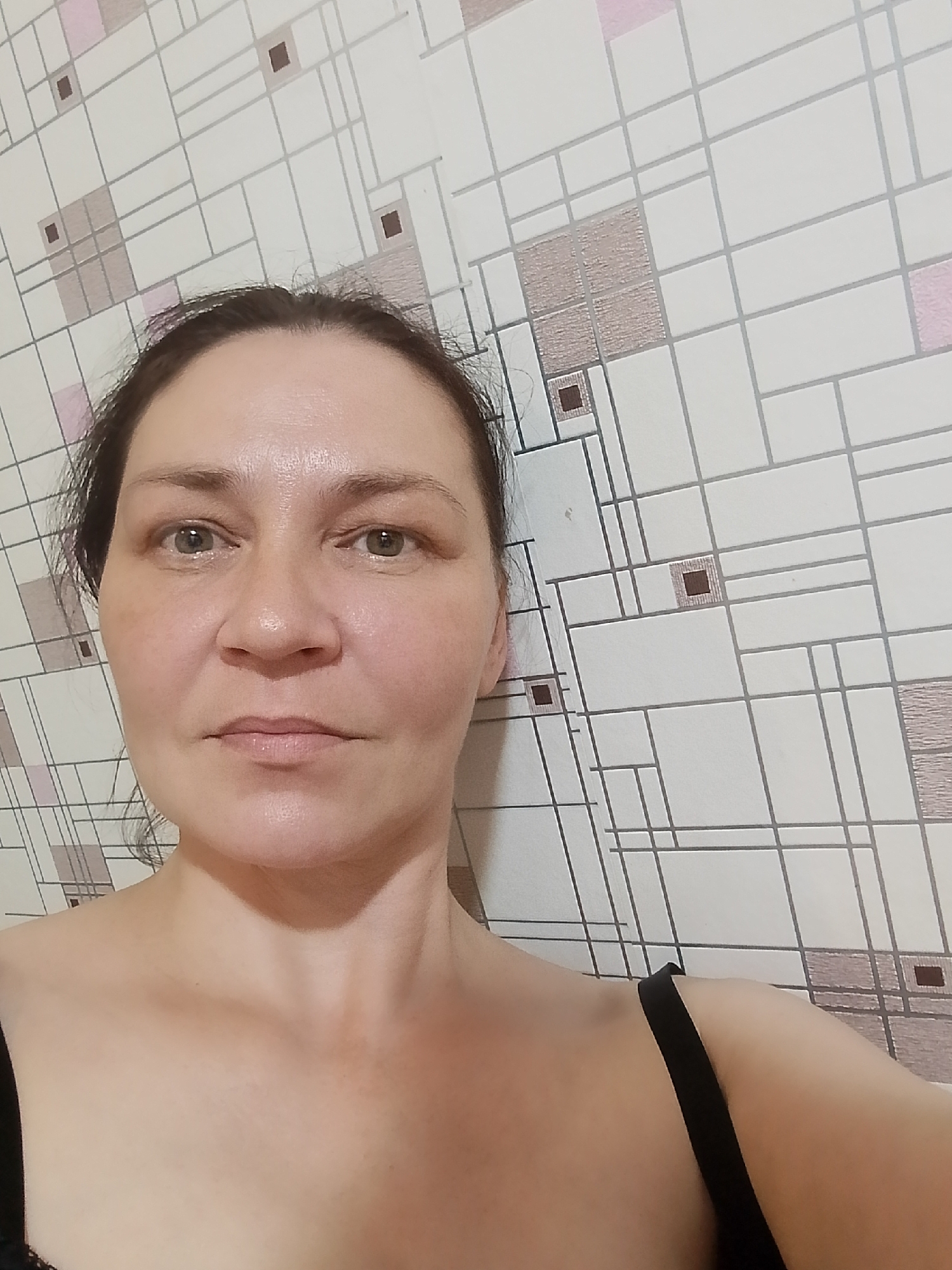A woman is photographed from the chest up, facing forward, with her head slightly tilted to her left. She has dark hair pulled back, light-colored eyes, and is wearing a dark garment, possibly a tank top or bra. The background is a wall with a geometric pattern of white squares and rectangles, interspersed with pink and brown squares. The lighting is even, suggesting an indoor setting. The image appears to be a selfie.