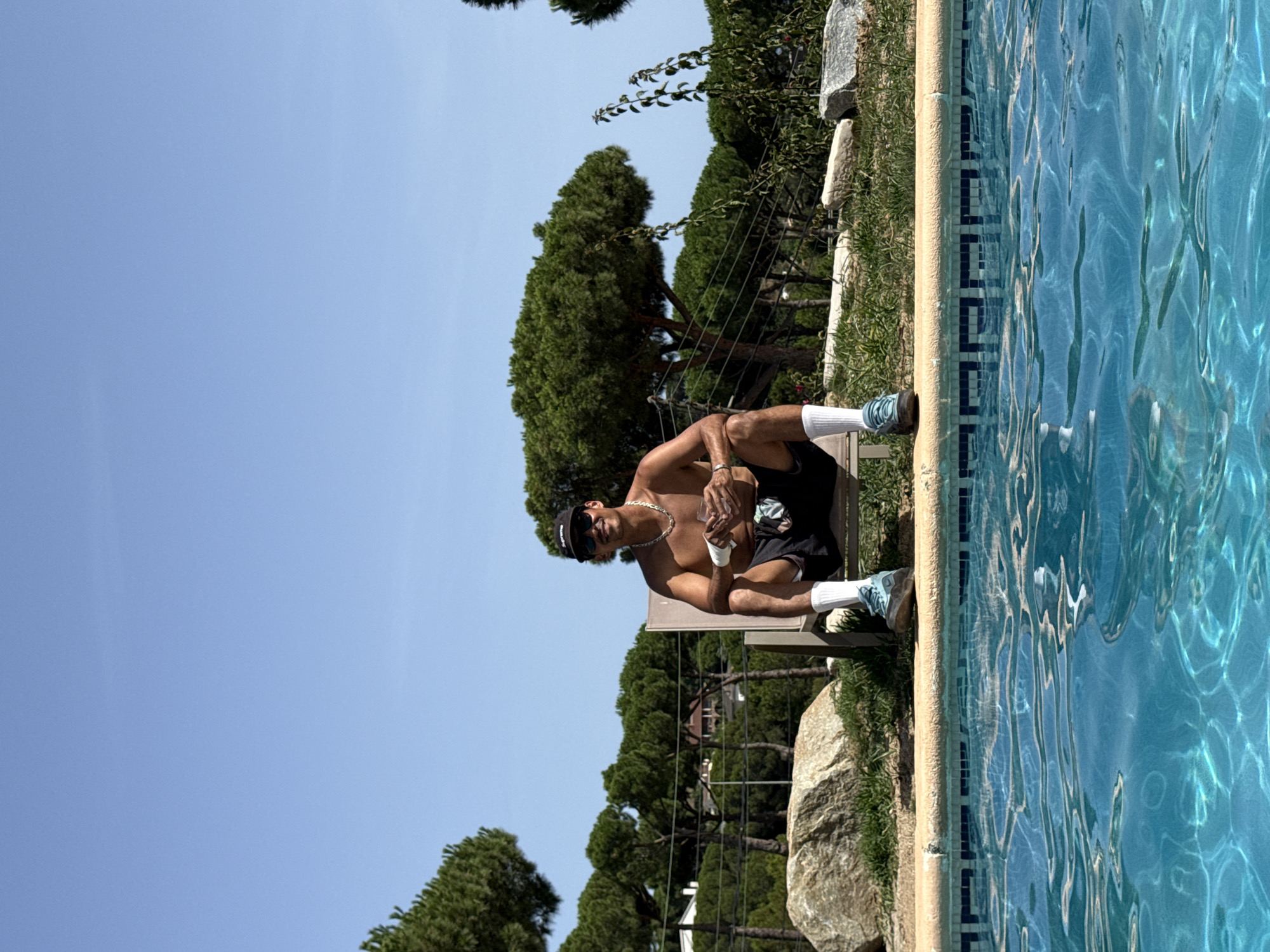 A shirtless man wearing black shorts, white socks, and blue sneakers sits on a lounge chair by a swimming pool. He is wearing a black baseball cap and sunglasses, and has a gold chain around his neck. He holds a clear glass with a beverage in his right hand. The man is outdoors with lush green trees and a clear blue sky in the background. Large rocks and grass surround the pool area. The scene appears to be a private residence in Sant Vicenç de Montalt, Spain, likely a backyard with a pool.