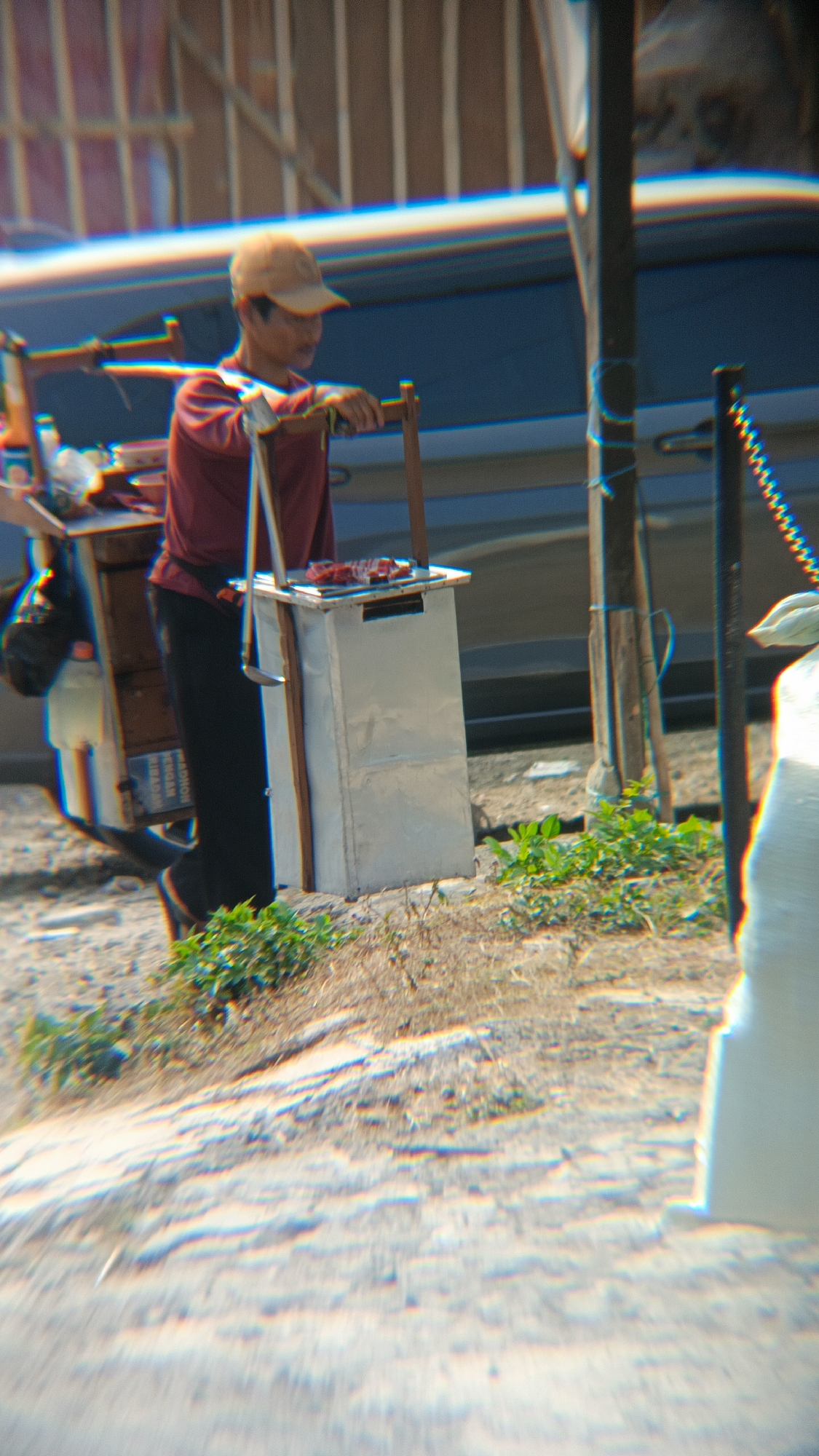 A person wearing a reddish-brown long-sleeved shirt and black pants, topped with a light brown baseball cap, stands next to a food cart. The cart, a mobile unit, has a metal structure and appears to be selling food, as raw meat is visible on a tray at the top. The person is holding onto a wooden pole that is part of the cart's support system. The background features a body of water with a blue hue, and vertical structures that suggest a building or a fence. The ground is a mix of dirt and sparse vegetation. The scene is set in Margajaya, Indonesia.
