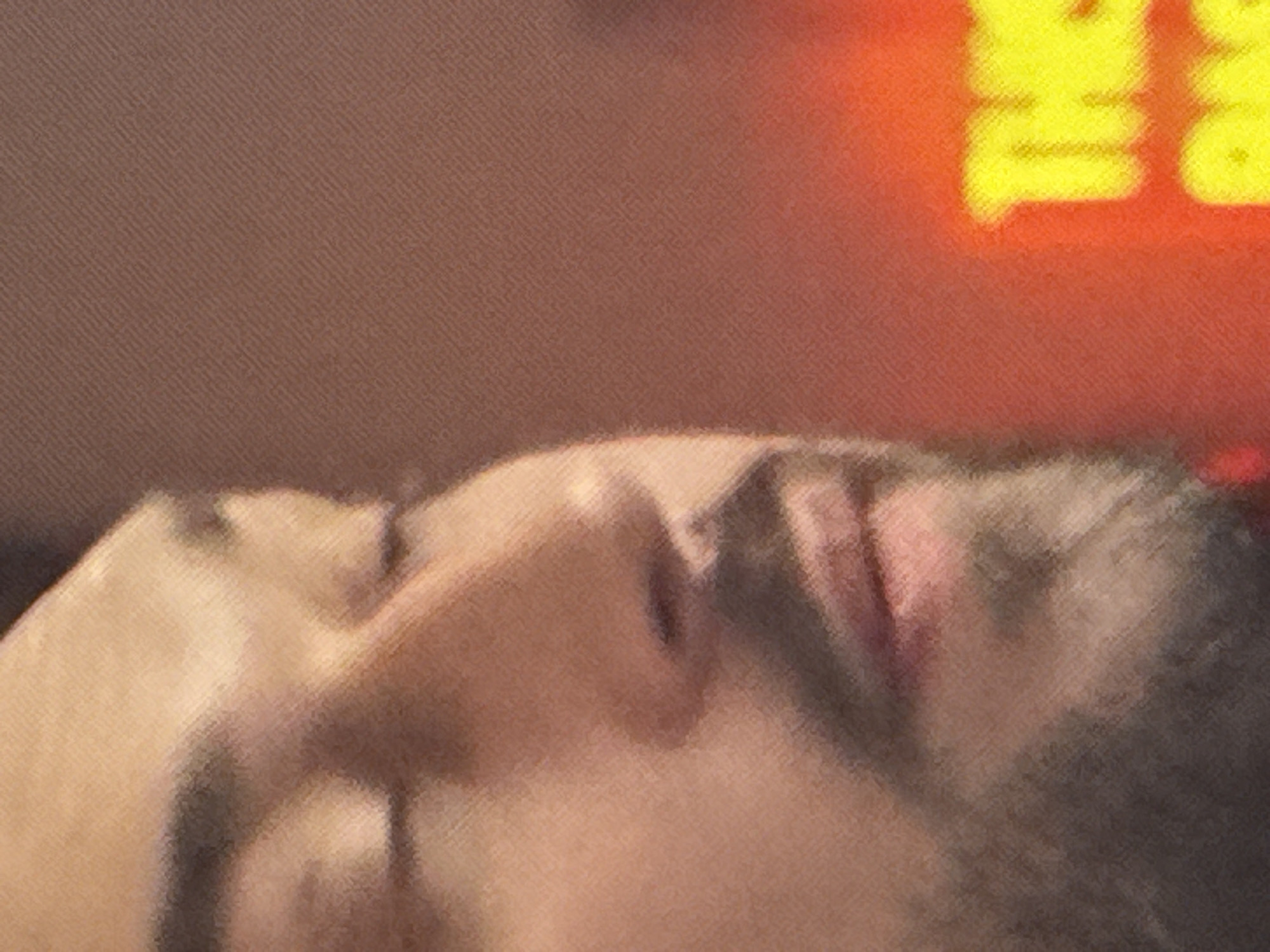 This is a close-up, side profile, low-angle shot of a man's face. He is looking down and to his right, with his eyes closed and his mouth slightly open. The man has dark hair and a beard. The background is a blurred red and orange gradient, with a portion of yellow text that appears to be part of a sign. The image is grainy and lacks specific details that would indicate a location in Saratov, Russia.