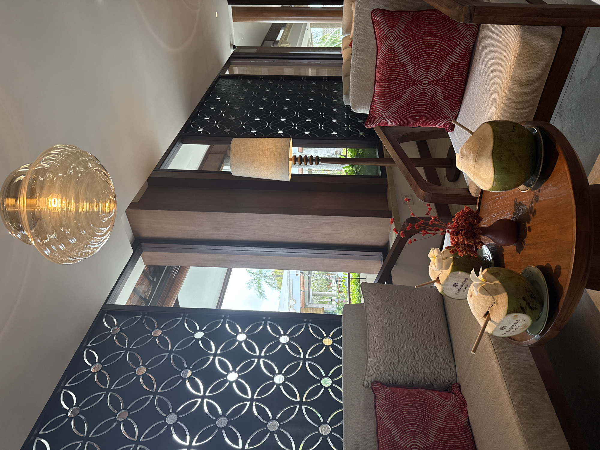 In Kuta Selatan, Indonesia, a seating area within a resort or hotel features a coffee table with two coconuts, each with a straw and a white flower. The coconuts are on coasters, and a small vase with dried red flowers sits nearby. The table is made of dark wood. Two chairs with light-colored cushions and red patterned throw pillows are positioned around the table. Behind the chairs, a decorative patterned screen with a geometric design divides the space. A floor lamp with a beige shade stands to the left, casting a warm light. The background shows a large window with a view of outdoor greenery and architectural elements. A large, ornate glass pendant light hangs from the ceiling. The overall ambiance is relaxed and tropical.