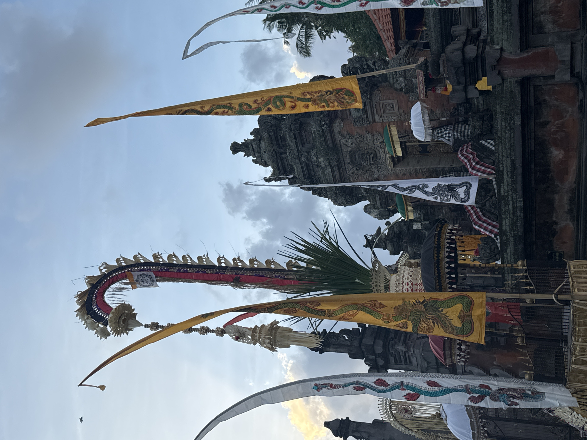A temple complex adorned with decorative banners and flags is pictured under a cloudy sky in Blahbatuh, Indonesia. The main structure is a multi-tiered stone building with intricate carvings and a prominent entrance. Several tall, narrow flags are planted in front of the temple, some featuring dragon motifs in yellow and white. A large, arched red and black decorative element, possibly a ceremonial banner, rises from the temple grounds. Other adornments include white, cone-shaped objects hanging from poles and several decorative umbrellas. The scene is set during daylight hours, with natural light illuminating the structures. No people are visible in the frame.