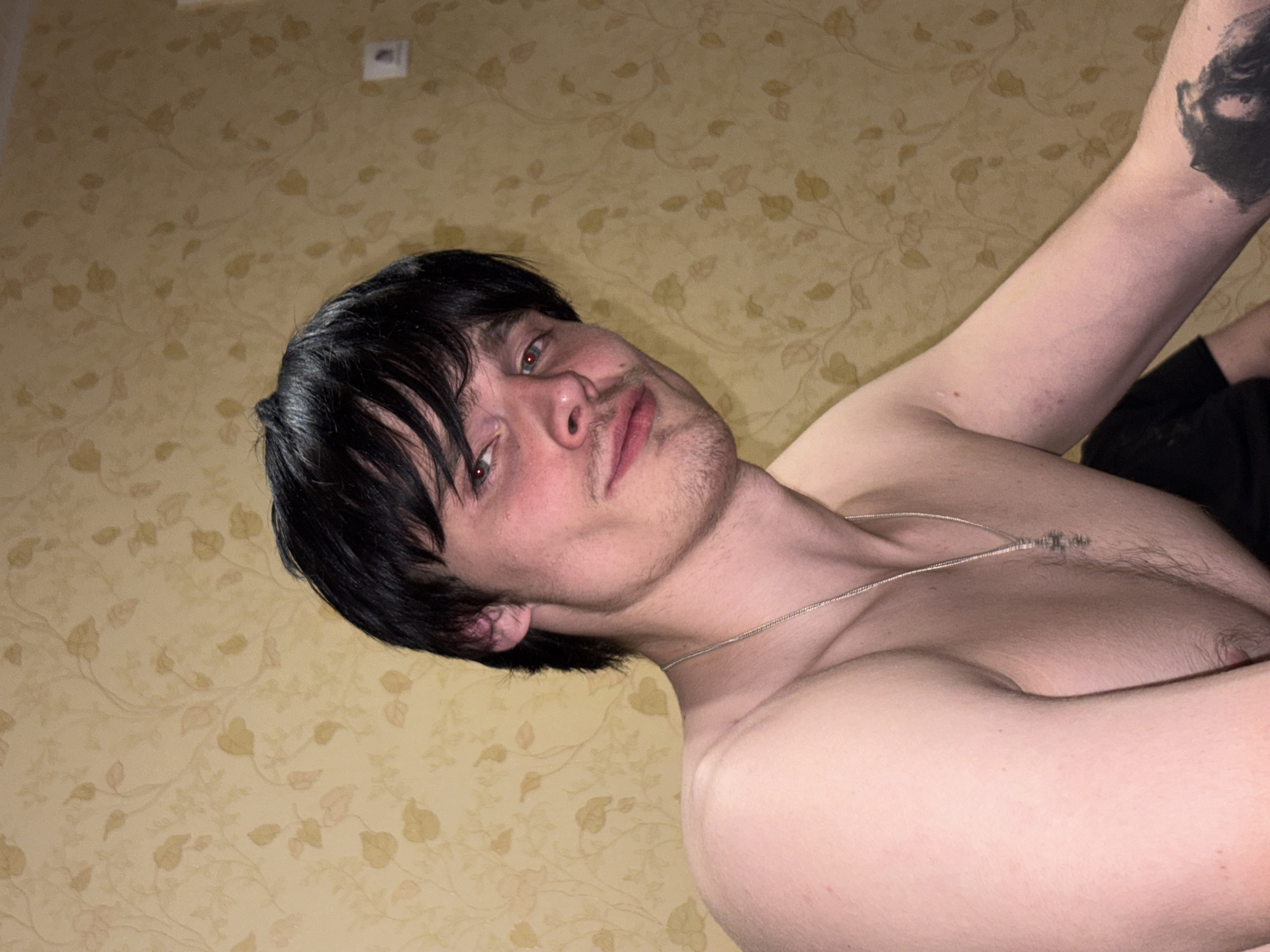 A young man is depicted from the chest up, looking directly into the camera. He has dark, medium-length hair with bangs, light-colored eyes, and light stubble on his face. He is shirtless, revealing his upper torso and arms, and wears a thin silver chain necklace. A dark, abstract tattoo, which appears to include an eye, is visible on his upper left arm. His skin is pale, and a small mark is noticeable on his right forearm.
The background features light beige or yellow wallpaper adorned with a subtle, lighter-toned floral or leaf pattern. A small, light-colored rectangular object, possibly a light switch or outlet, is visible on the wall in the upper left corner. The image appears to be taken indoors, under artificial lighting, and is angled as if the subject is lying down or holding the camera tilted. The man's expression is neutral and direct. No text is visible in the image. The setting is an indoor residential space, with the provided context indicating the location as Chaikovsky, Russia.