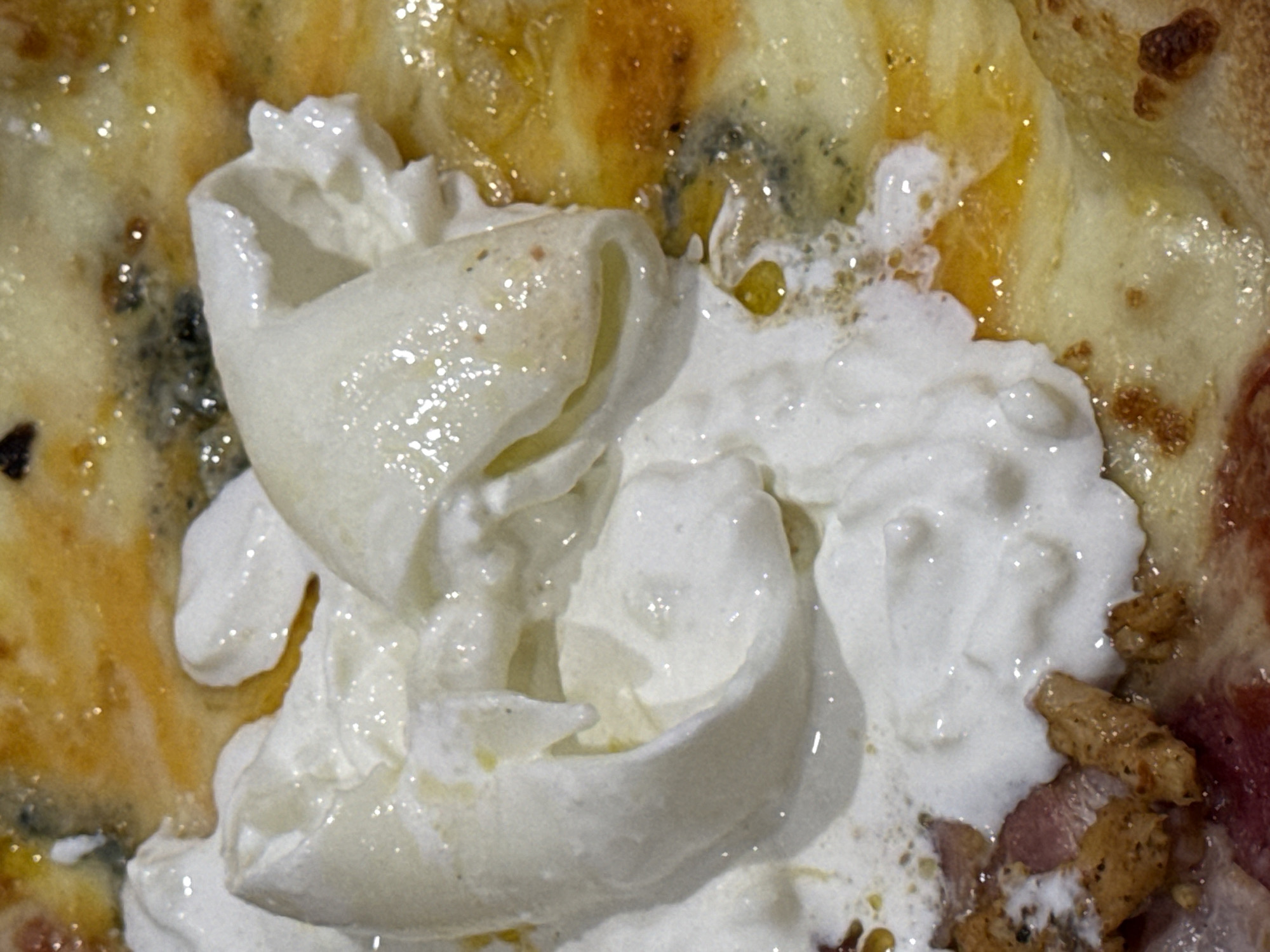 A close-up shot of a pizza slice is captured, with toppings including cheese, cream, and meat. The cheese appears melted and slightly browned in areas. A dollop of creamy white cheese is positioned centrally, with a distinct curled shape. The meat appears cooked and fragmented. The overall texture is glossy, suggesting some oil content. The image does not offer any information to determine the location is in the city of Đà Nẵng, Vietnam.