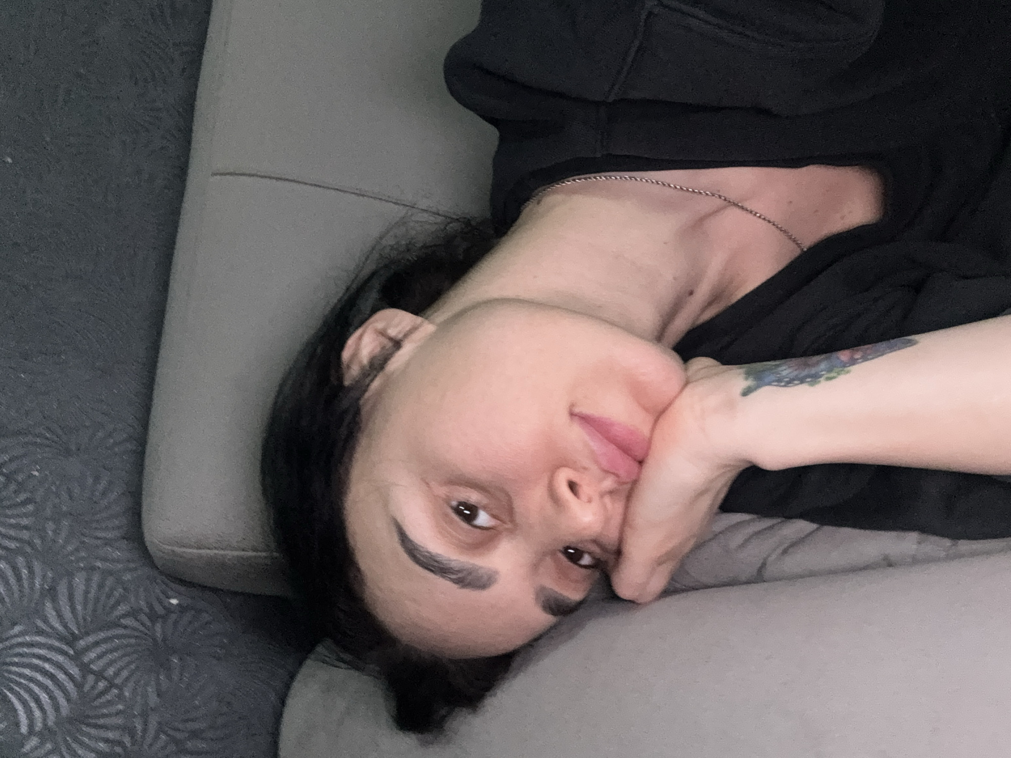 An indoor, eye-level shot features a person lying on their side on what appears to be a light grey sofa or bed. The person, an adult with dark hair and prominent eyebrows, is looking directly at the camera with a slight, relaxed smile. Their head is tilted, and they are resting their chin on their left hand, which is supporting their head. A colorful tattoo, possibly floral, is visible on their inner left forearm. They are wearing a dark, possibly black, long-sleeved top or hoodie, and a thin silver-colored chain necklace is visible around their neck. The background consists of the grey upholstered furniture, with one section showing a subtle dark grey pattern resembling stylized leaves or waves. The lighting is soft and even, suggesting an indoor setting without strong natural light cues. No text is visible in the image.