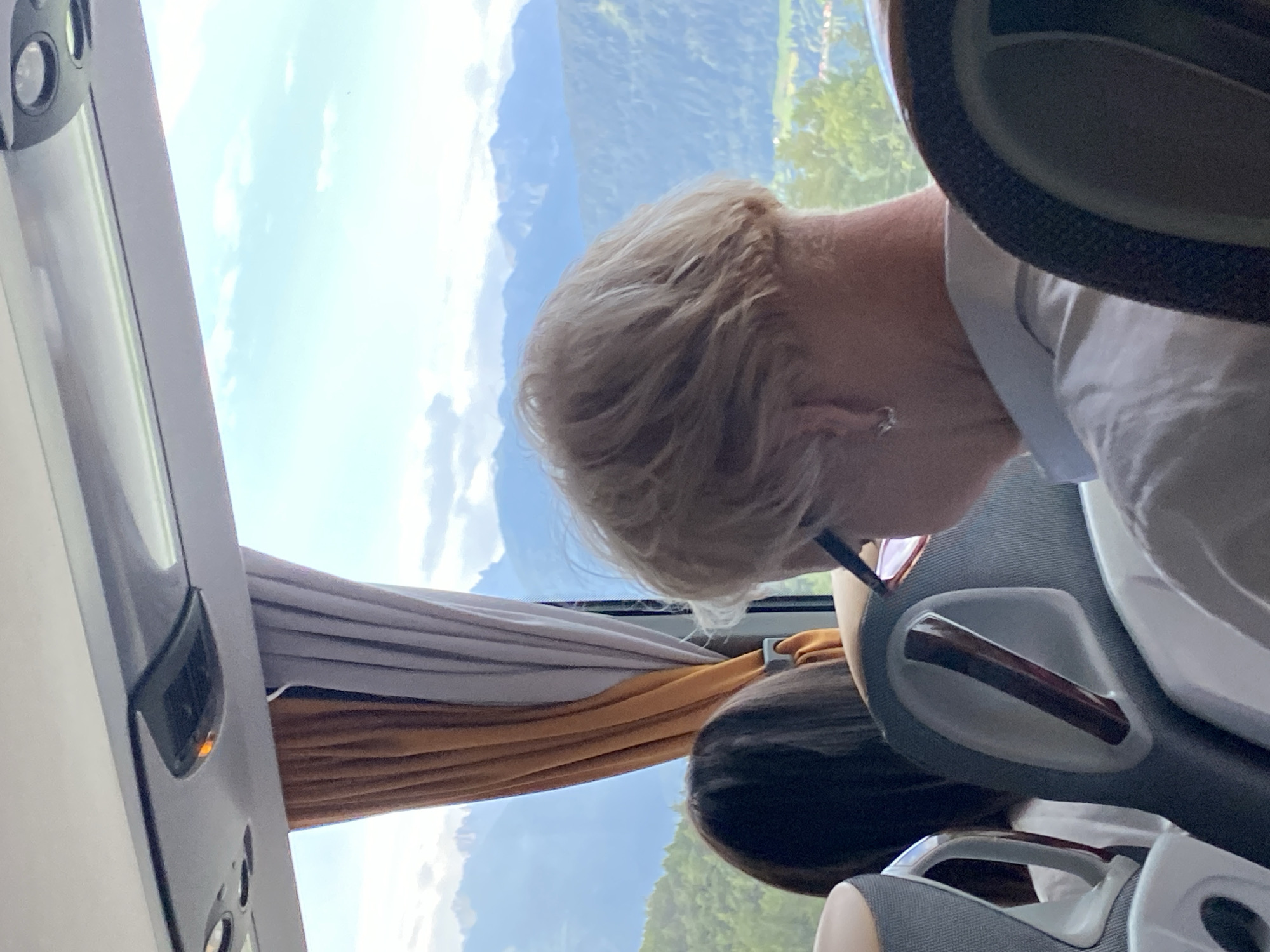 The image shows the interior of a bus, with passengers seated and looking out the window at a mountainous landscape. The window is partially covered by orange and grey curtains. In the foreground, the back of a person's head with short blonde hair is visible, wearing a white shirt and glasses. To the left, another passenger's dark hair is visible. The scene appears to be set in the Austrian Alps, specifically in or near Reith bei Seefeld, given the prominent mountain range in the background. The lighting suggests it is daytime, with the sky visible through the window.