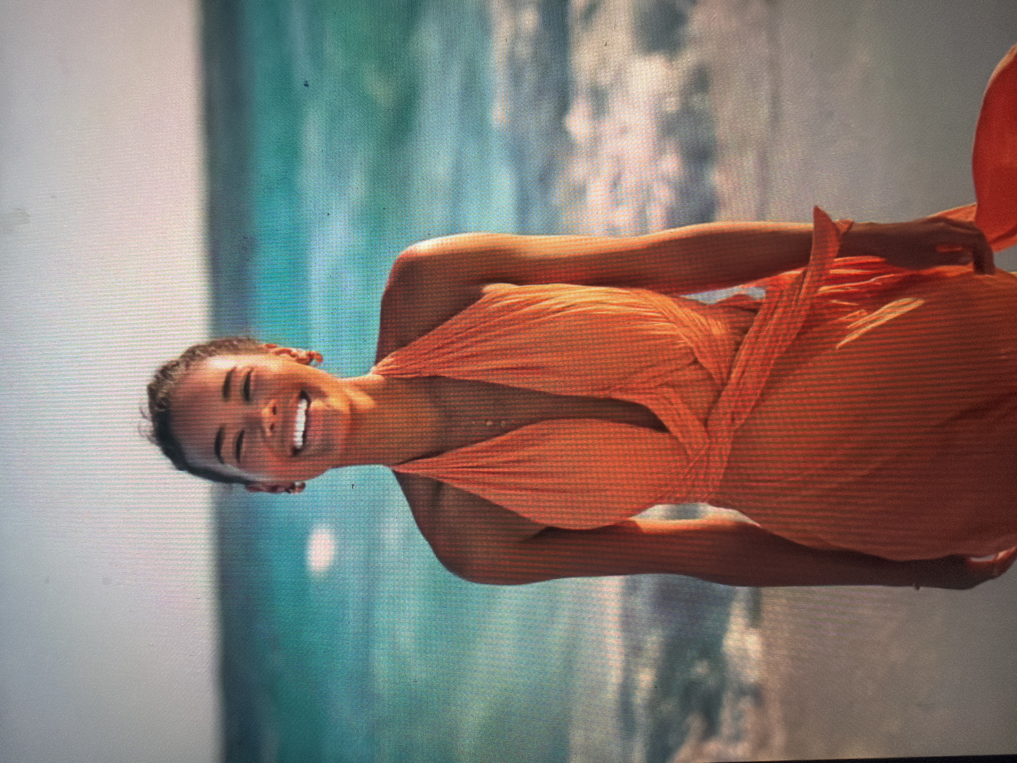 A young woman is depicted standing on a sandy beach. She wears a vibrant orange halter-neck dress or top with a deep V-neck and a wrap-around waist detail. Her dark hair is styled away from her face, and she presents a wide, open-mouthed smile, revealing her teeth. Small earrings are visible. The background features a clear turquoise ocean with white waves, extending to a pale sky at the horizon line. The immediate foreground shows the sandy beach. The image quality reveals visible screen pixelation and minor surface smudges, indicative of a photograph taken of a digital display. The depicted coastal environment is geographically inconsistent with Shumen, Bulgaria, which is an inland city.