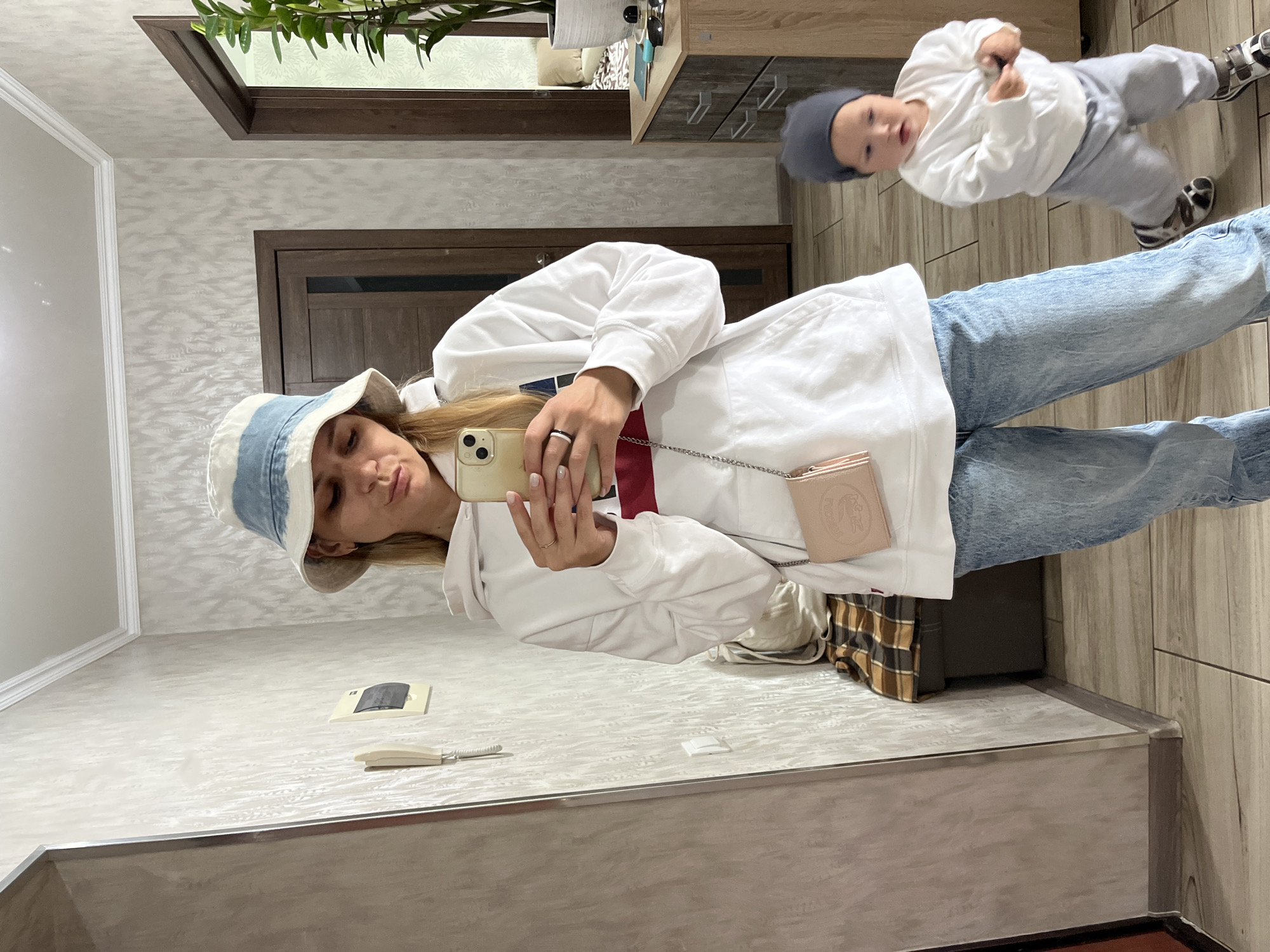 A blonde woman wearing a white and blue bucket hat, a white hoodie, and light-wash jeans takes a mirror selfie in a hallway. She is holding a smartphone in her right hand and a small pink purse with a chain strap is visible on her left side. In the reflection of the mirror, a toddler wearing a gray beanie, a white sweatshirt, and gray pants is in motion, appearing to be walking or running. The hallway features textured wallpaper, a wooden door, a bench with a plaid cushion, and a dresser with plants and sunglasses on top. The floor is tiled with a wood-grain pattern.