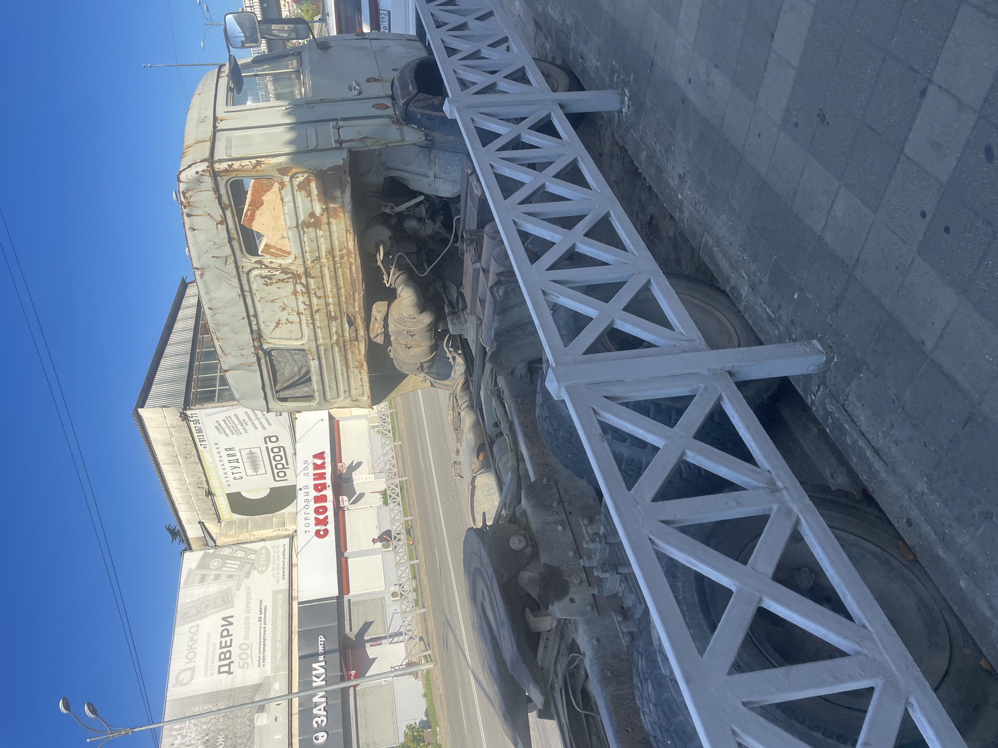 In Sochi, Russia, a weathered, light-colored truck cab rests on a sidewalk, its rear chassis extending onto the road. The cab shows significant rust and damage, with a broken window revealing exposed interior components. The truck is positioned behind a white, diamond-patterned fence. In the background, several businesses are visible, including "ЛЮККА ДВЕРИ" (Lyukka Doors), "Студия Город" (Studio Gorod), and "ТОРГОВЫЙ ДОМ С КОВЯНКА" (Trading House Skovyanka). A lone individual walks across the street in front of these establishments. The sky above is clear and blue, and the scene is illuminated by bright daylight, suggesting it is daytime in a coastal urban setting.