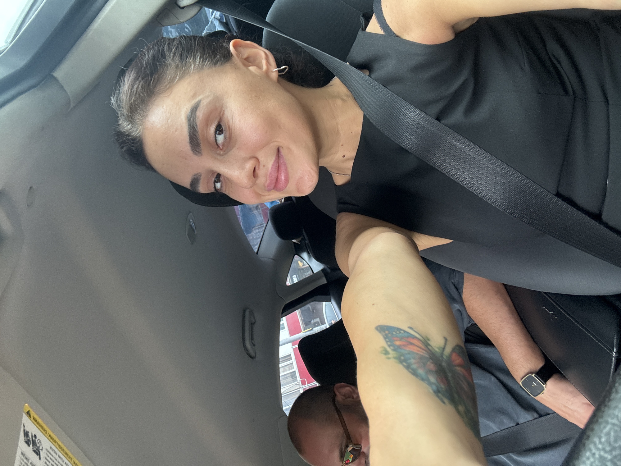 A woman with dark hair, wearing a black dress and a seatbelt, takes a selfie in a car. She has a butterfly tattoo on her arm. A man with glasses is visible in the passenger seat. The car appears to be driving in a city environment with buildings visible outside the windows.
