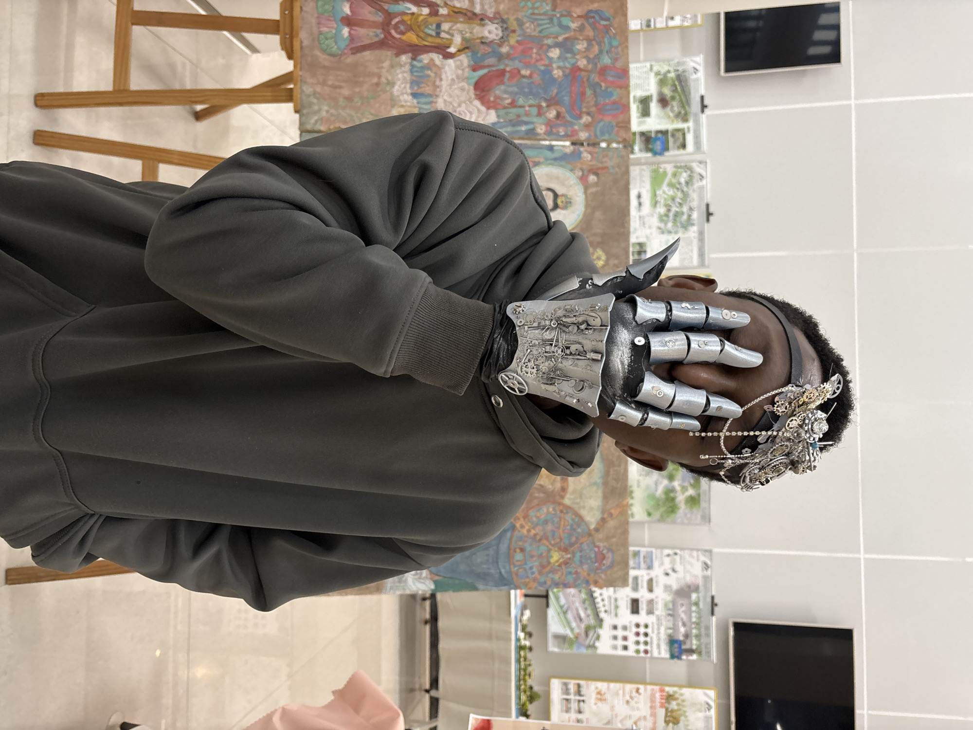 A person wearing a gray hoodie, a metallic prosthetic hand, and a decorative headpiece is shown in a medium shot. The person is partially obscuring their face with their prosthetic hand. The background includes artwork on easels, architectural drawings displayed on a wall, and a white-tiled floor. The location is identified as Jiangjin, China.