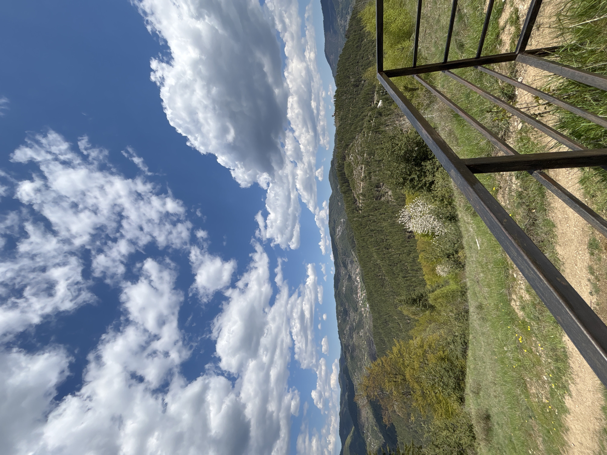 The image presents a landscape view from an elevated position, potentially from a man-made structure, which is indicated by a metal railing in the right-hand side of the frame. The perspective appears tilted, rotating the scene 90 degrees counter-clockwise. The landscape consists of a mountain range covered in dense forest. In the far background, the sky features cumulus clouds against a clear blue background. The foreground consists of grassland with visible yellow wildflowers, and some scattered trees. It is not possible to ascertain the location in the city of Gradat, Bulgaria. No people, subjects, objects, actions, or interactions are clearly visible.