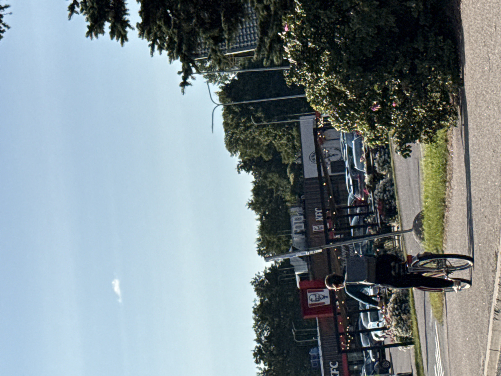 A person rides a bicycle away from the camera, down a paved road in Tallinn, Estonia. The cyclist, wearing dark pants and a dark long-sleeved shirt with a grey backpack, is moving from left to right in the frame. The bicycle is dark-colored with red accents on the wheels and frame. In the background, a row of buildings with prominent KFC signage is visible, along with parked cars. The environment appears to be an urban or suburban area with trees and streetlights. The sky is clear and blue with a small cloud.