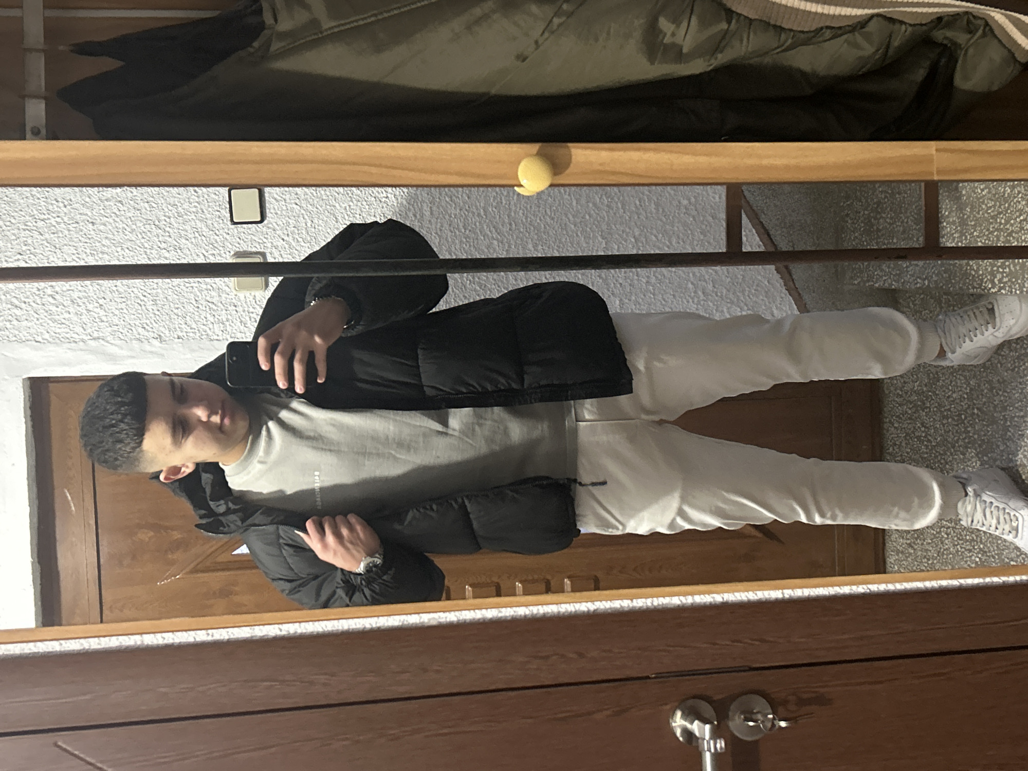 A young man is taking a selfie in a mirror. He is wearing a grey t-shirt, white sweatpants, white sneakers, and a black puffer jacket. The mirror is in a room with wooden doors and trim. There is a shelf above the mirror with a green jacket on it. The wall is white with a textured surface. A yellow object is sitting on the frame of the mirror. The location is likely indoors but the context doesn't indicate that it is in Zlatograd, Bulgaria.