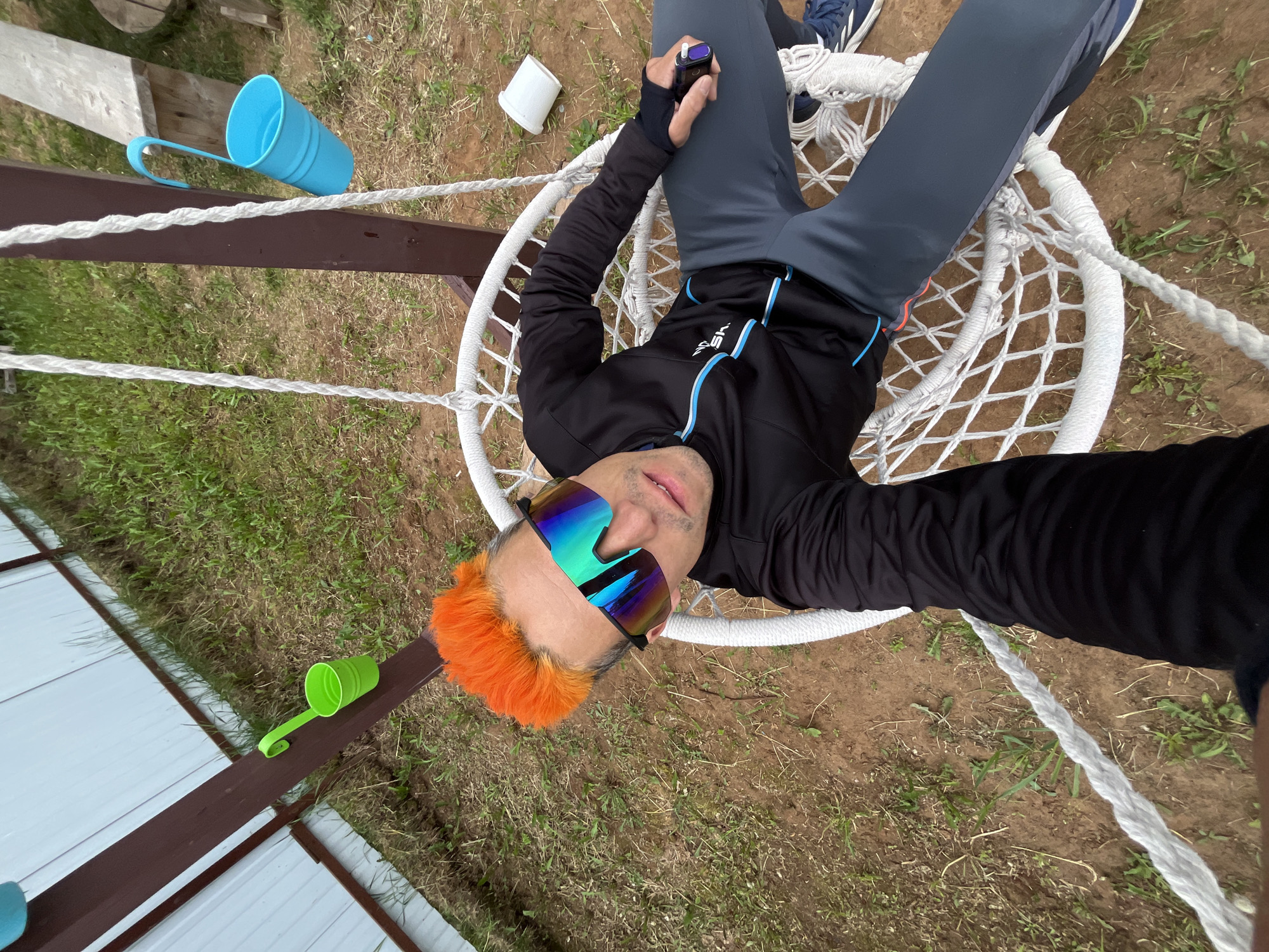 An overhead selfie captures a person relaxing in a white, circular woven hanging chair outdoors. The subject, likely male, has distinct short, bright orange hair and wears large, reflective, multi-colored sunglasses. They are dressed in a black long-sleeved athletic jacket with blue accents and the visible brand name "HEAD" on the chest, dark grey athletic pants, and blue sneakers. In their right hand, they hold a small, dark electronic device with a white tip, possibly a vaping device.
The hanging chair is positioned over a ground that consists mostly of bare brown earth with scattered patches of green grass and weeds. A dark brown wooden beam, appearing to be part of a fence or support structure, traverses the frame. Hanging from this beam are two plastic mugs: one light blue and one lime green, both with handles. A small white cylindrical object lies on the ground near the person's left arm. In the background on the left, a white corrugated metal wall or fence is partially visible.
The scene depicts a casual outdoor setting, suggesting a backyard or rural space, under natural daylight. The person appears relaxed while taking the photo. The setting for this media file is Chaikovsky, Russia.