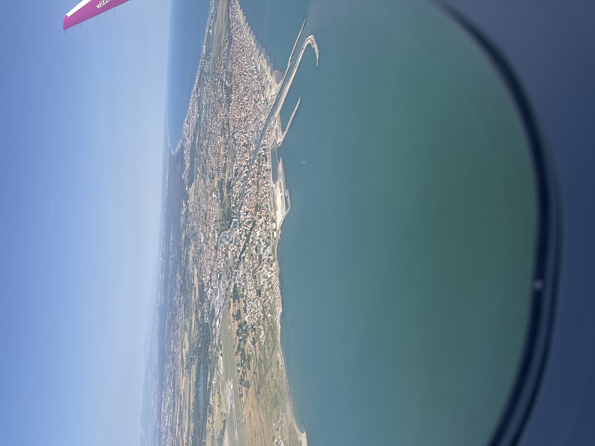 The image shows an aerial view of Fiumicino, Italy, captured from the window of an aircraft, likely a commercial plane, with a portion of a pink Wizz Air plane wing visible in the upper left. The scene depicts a coastal cityscape meeting the Tyrrhenian Sea, characterized by a dense urban area stretching along the coastline. The image also reveals an airport with visible runways and adjacent agricultural land. The city has a prominent harbor with curved breakwaters. The water is a dark turquoise color. The photo appears to have been taken during the daytime under sunny conditions.