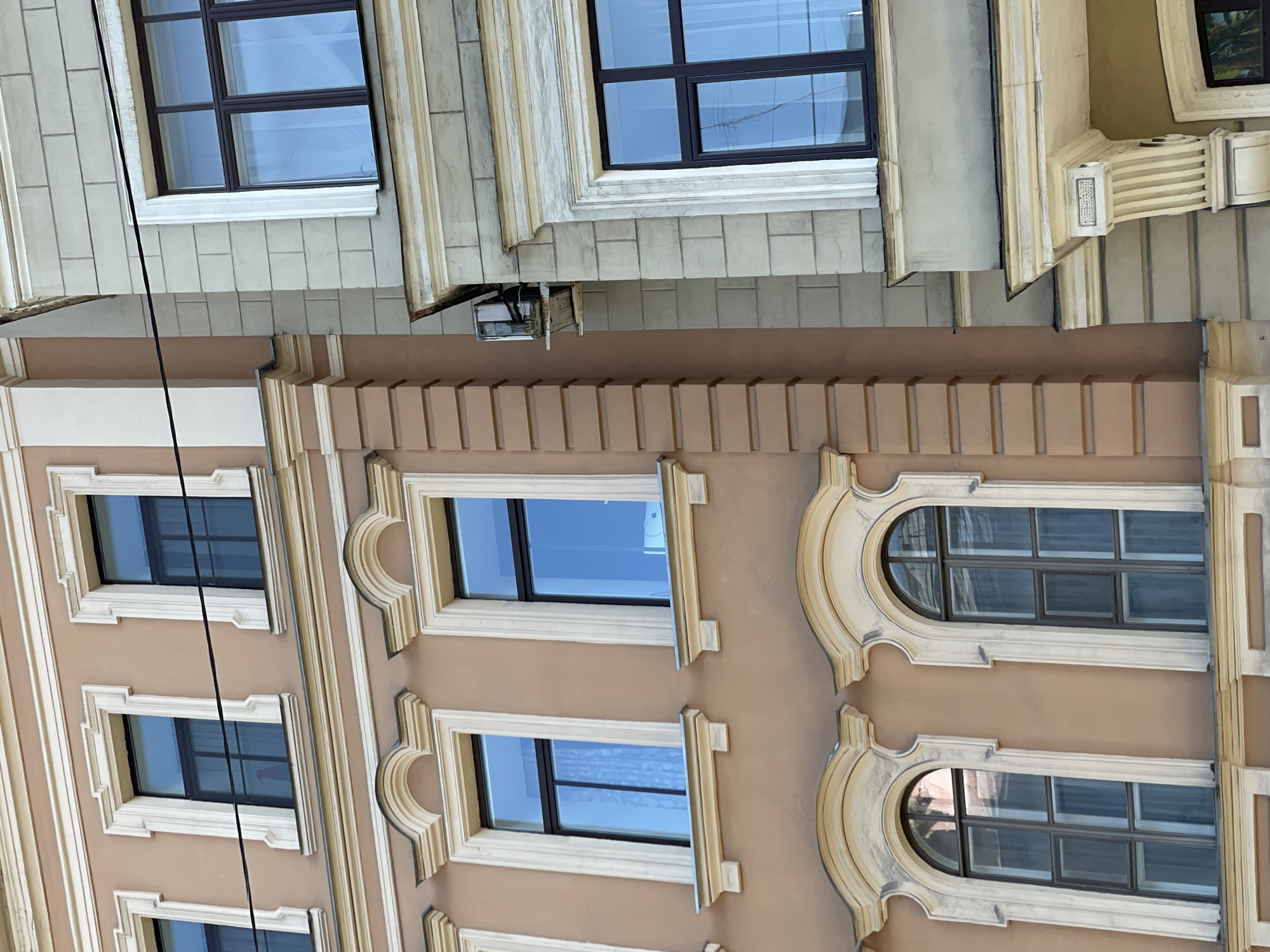 The image shows a portion of two buildings in what is likely Saint Petersburg, Russia, given the architectural style. The buildings are constructed with a classical design, featuring decorative windows and facades. The upper building is grey with rectangular windows, while the lower building is painted tan with both rectangular and rounded windows. A black cable runs vertically along the facade. The sky is visible in the windows.