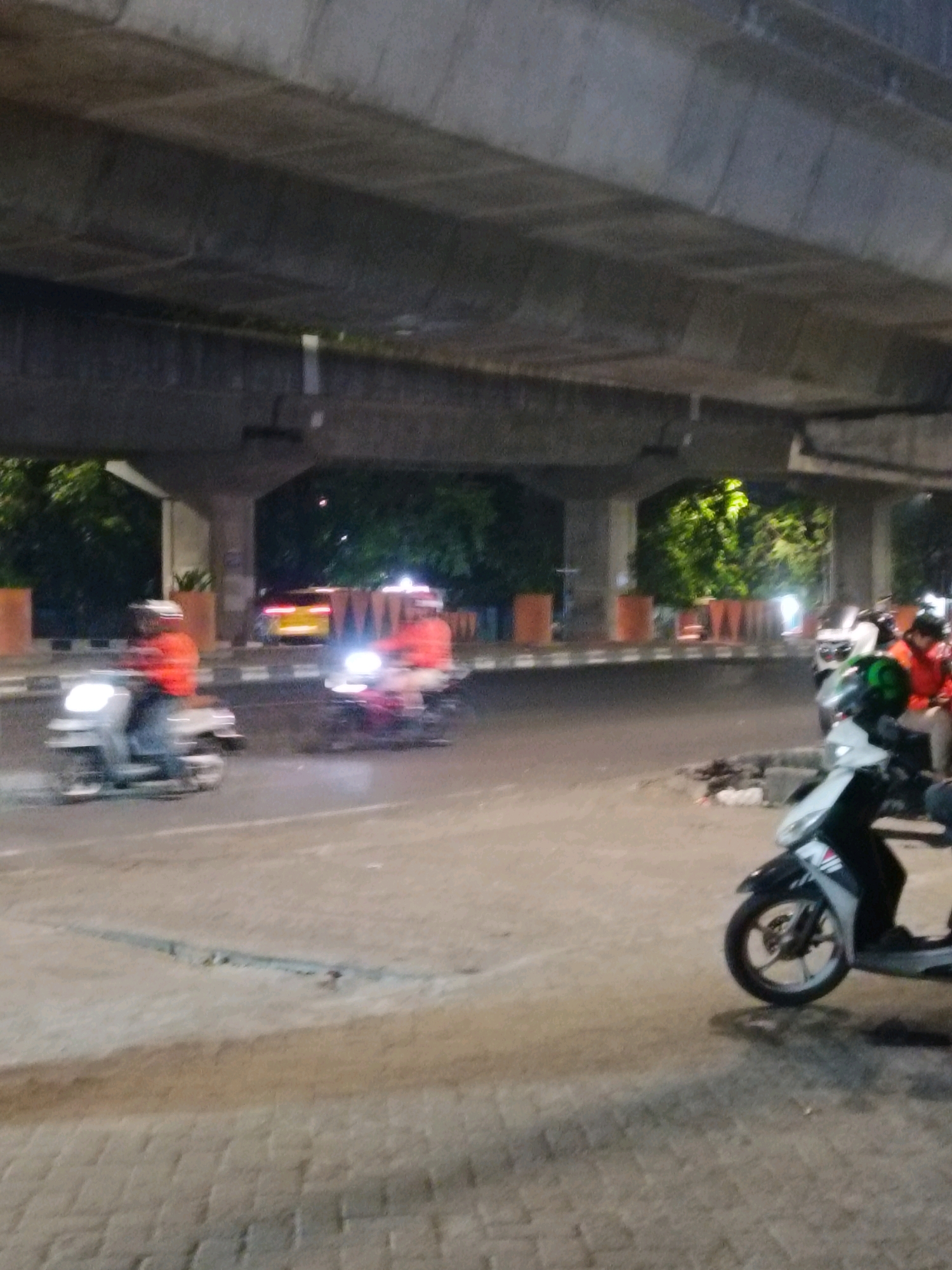 An outdoor, medium shot captures a street scene at night, likely in Makassar, Indonesia, under a concrete overpass. The image shows several people on motorcycles, with motion blur indicating movement. In the foreground, a person on a white scooter is partially visible. Further down the road, two individuals in orange jackets are riding motorcycles, their headlights illuminated. A yellow car is visible in the background. The environment includes a paved road, a sidewalk with brick paving, and planters lining the street. Trees and foliage are visible behind the planters, illuminated by artificial light. The overall impression is one of a busy urban thoroughfare at dusk or night.