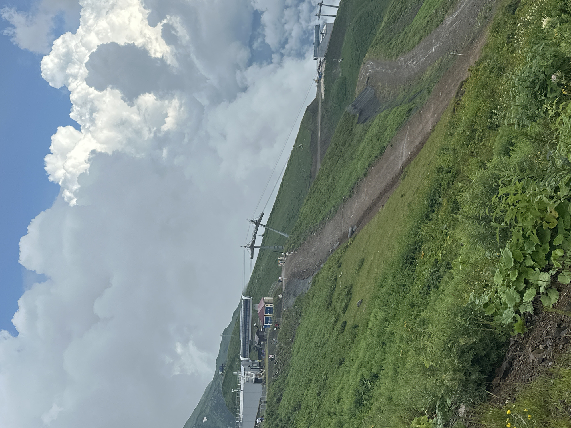 This image captures a scenic mountain landscape in Estosadok, Russia, with a ski lift system prominent in the mid-ground. Lush green grass covers the sloping hills, interspersed with some darker, bare earth patches, suggesting trails or erosion. The sky is a dramatic mix of bright blue and large, billowing white clouds.
In the foreground, dense green vegetation, including large-leafed plants and smaller wildflowers, grows on a rocky incline. A dirt path winds its way up the mountainside.
Further up the slope, a complex ski lift structure is visible. This includes a station with a white building and a covered conveyor belt, from which ski lift cables extend upwards. Several people, appearing as small figures in the distance, are present near the ski lift station. A small, colorful building with a red roof and blue walls sits adjacent to the main station.
A ski lift tower, a metal pole with supporting cables, stands on the slope. In the distance, another ski lift station or structure can be seen on a higher ridge. The overall impression is a mountain resort area on a cloudy but visually striking day.
