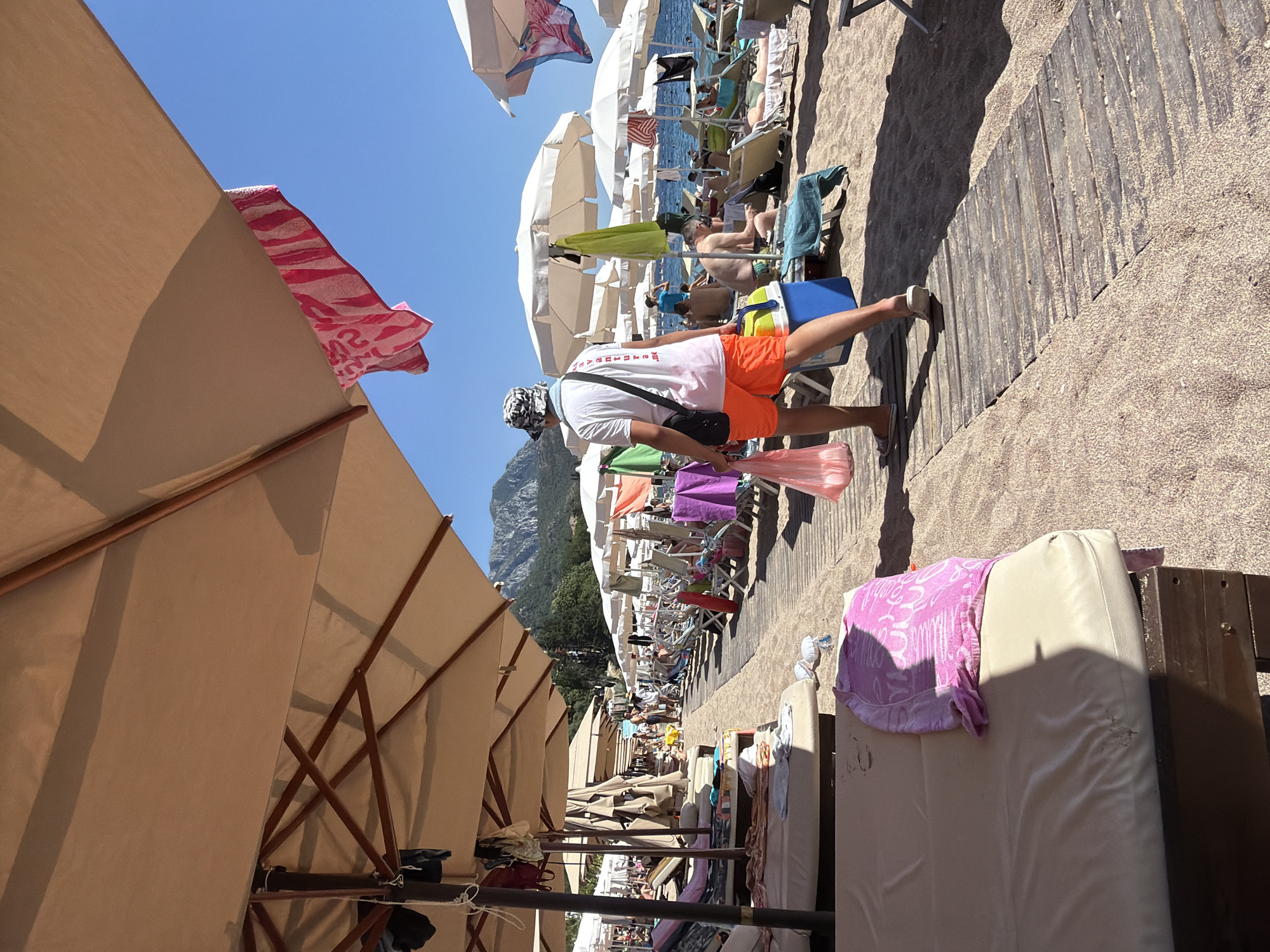 A man walks on a wooden boardwalk on a crowded beach. He is wearing orange shorts, a white t-shirt, and a patterned hat. He carries a pink plastic bag in his left hand and a blue and yellow cooler in his right. Several beach umbrellas and lounge chairs are visible, with people sunbathing and sitting under the umbrellas. The beach is sandy, and the sea is in the background. Mountains are visible in the distance, and the sky is clear and blue.