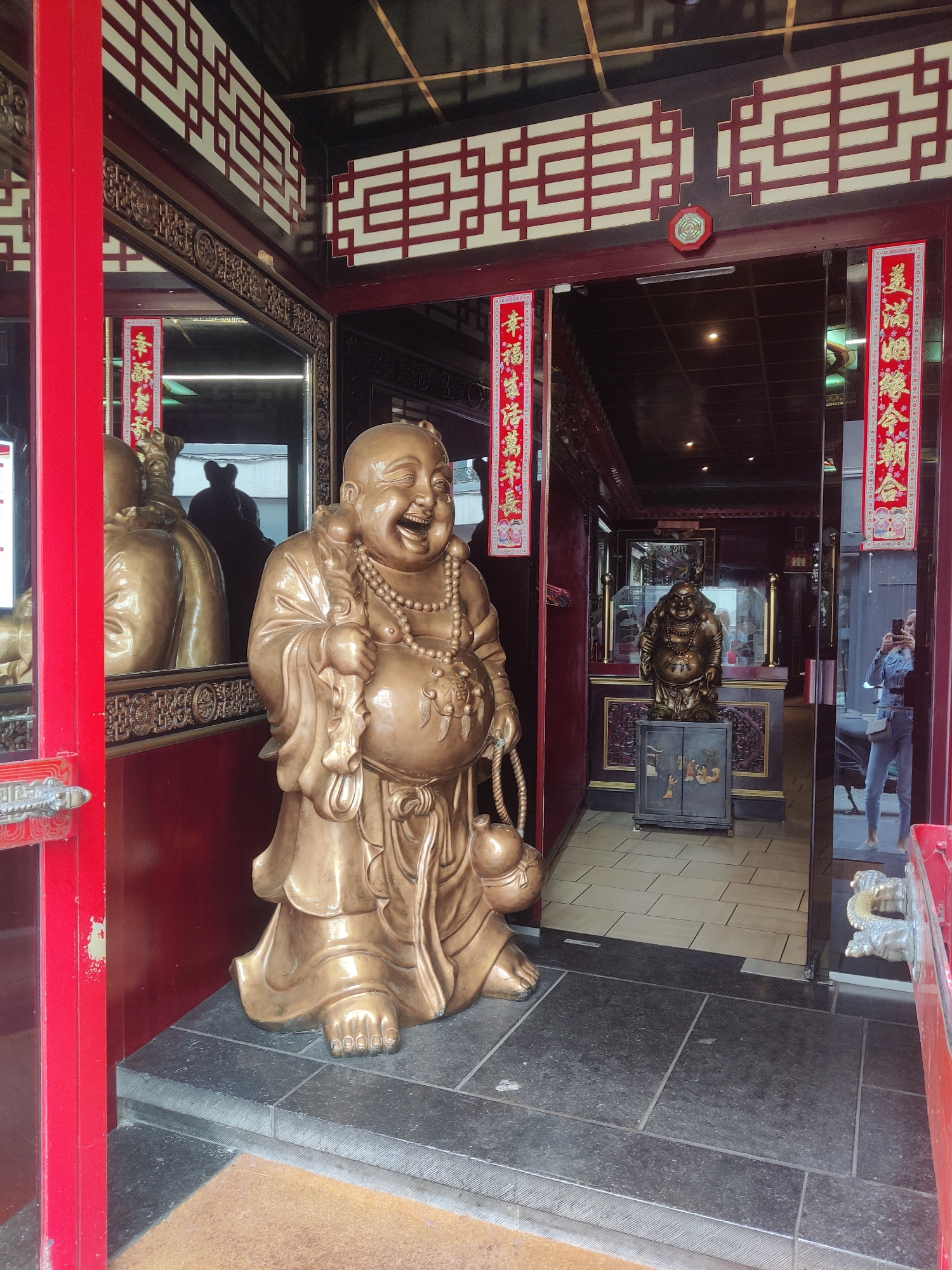The image shows the entrance to a Chinese restaurant in Liège, Belgium. Two large golden statues of a laughing Buddha are prominently displayed. The statue in the foreground is positioned on the left side of the entrance, and another statue is visible further inside the restaurant. Red banners with Chinese characters hang on either side of the entrance. The restaurant's interior features dark wood paneling and decorative screens. A person is visible taking a photo of themselves in the reflection of the glass door. The overall scene suggests a festive and welcoming atmosphere typical of a Chinese establishment.