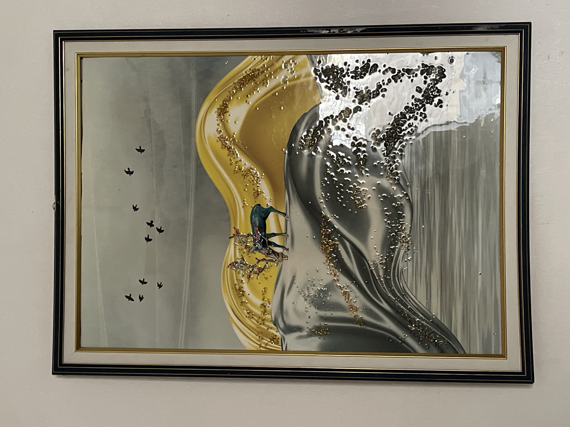 A framed piece of artwork hangs on a white wall. The artwork features a stylized abstract design with gold and silver colors prominently displayed. The gold and silver sections appear to flow and intertwine, with textured, raised details that add depth. On the left side, a flock of black birds is depicted flying upwards. In the center, a stylized bird with green and blue hues is perched on a branch with red berries. The frame is a dark color with a thinner gold accent. The surrounding wall is plain white. There is no information about Kuchingoro, Nigeria in this image.