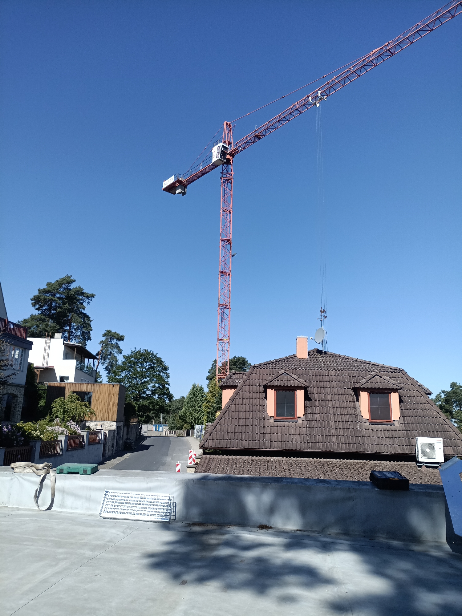 In Doksy, Czechia, a construction crane is prominent against a clear blue sky, its red lattice structure reaching high. The crane's jib extends over a residential area featuring houses with brown tiled roofs, some equipped with dormer windows and an air conditioning unit. In the foreground, a flat rooftop area is visible, with a grey membrane, a white grate, and a green tool case. Adjacent to the rooftop, a street curves uphill, lined with trees and houses. A portion of a multi-story building with balconies and stone detailing is also present on the left side of the image.