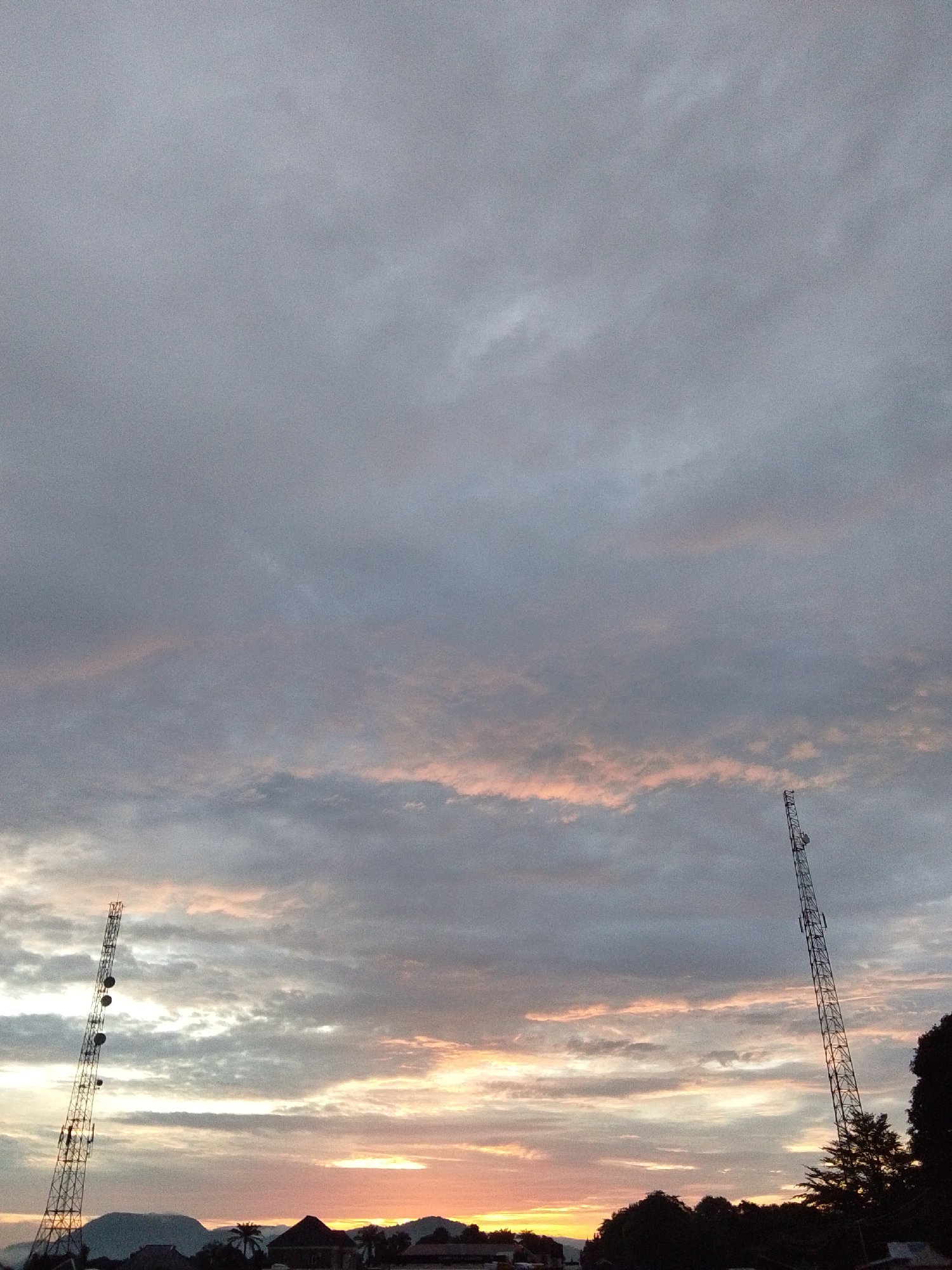 The image depicts a serene sunset scene in the city of Serti, Nigeria. The sky is filled with a dramatic display of clouds, ranging from deep gray to soft pink and orange hues, illuminated by the setting sun. Two tall communication towers stand prominently against the twilight sky, one on the left and another on the right, their metallic structures silhouetted. Below the towers, a residential area is visible, with rooftops and trees forming a dark, natural barrier. The overall impression is one of tranquility and natural beauty.