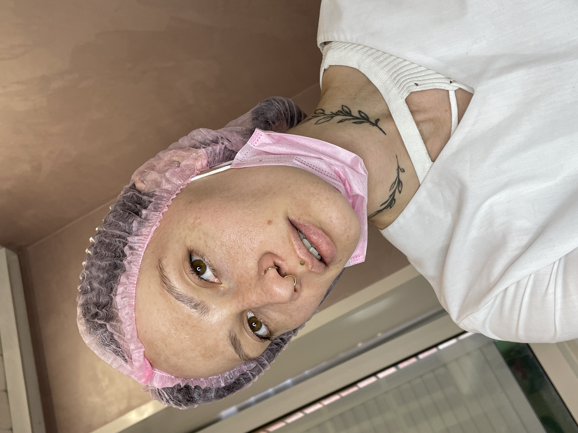 A woman is lying on her back, wearing a pink surgical mask, a hairnet, and a white garment. She has tattoos on her neck and piercings in her nose and upper lip. The background includes a pinkish-tan wall and a window. The photo may have been taken indoors in a medical or cosmetic setting. No information related to Izhevsk, Russia is visible.