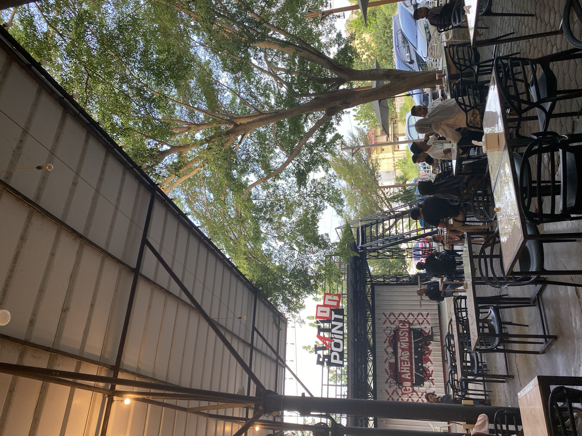 The image depicts an outdoor dining area, likely a cafe, situated in Lhokseumawe, Indonesia. The area is partially covered by a corrugated metal roof with exposed steel framing, under which warm-toned lighting is visible. Large trees with dense foliage provide additional shade overhead. Adjacent to the dining area is a wall or partition with signage that reads "ST4TION POINT" in vertically stacked letters. Below, a mural displays the text "GO AHEAD MUSIC" with an image resembling a vintage locomotive. Seated at tables are multiple individuals, some interacting with each other, some seemingly alone. The furniture consists of black plastic chairs and light-colored tables. Beyond the immediate area, cars are parked.