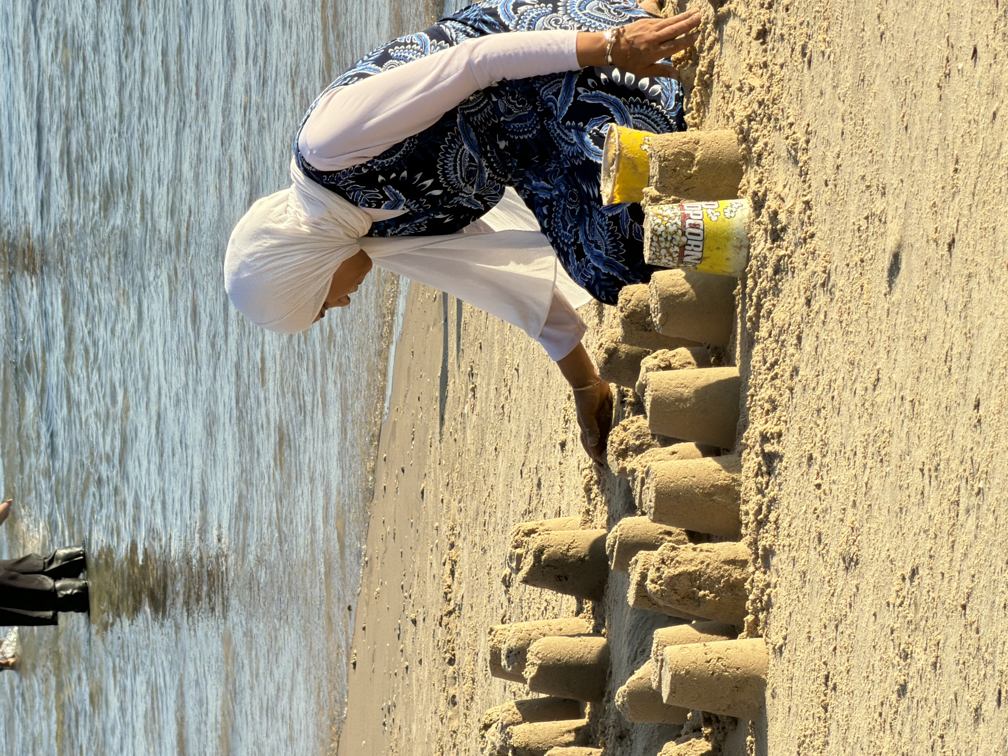 A woman wearing a white hijab and a blue and white patterned dress is kneeling on a sandy beach, building sandcastles. She is surrounded by numerous sandcastles of varying sizes. In front of her are two yellow containers, one appearing to be a popcorn bucket. The ocean is visible in the background. The scene appears to be taking place in Tel-Aviv, Israel, as indicated by the presence of people in the background wearing clothing that suggests a hot climate and the architecture of the buildings visible in the distance.