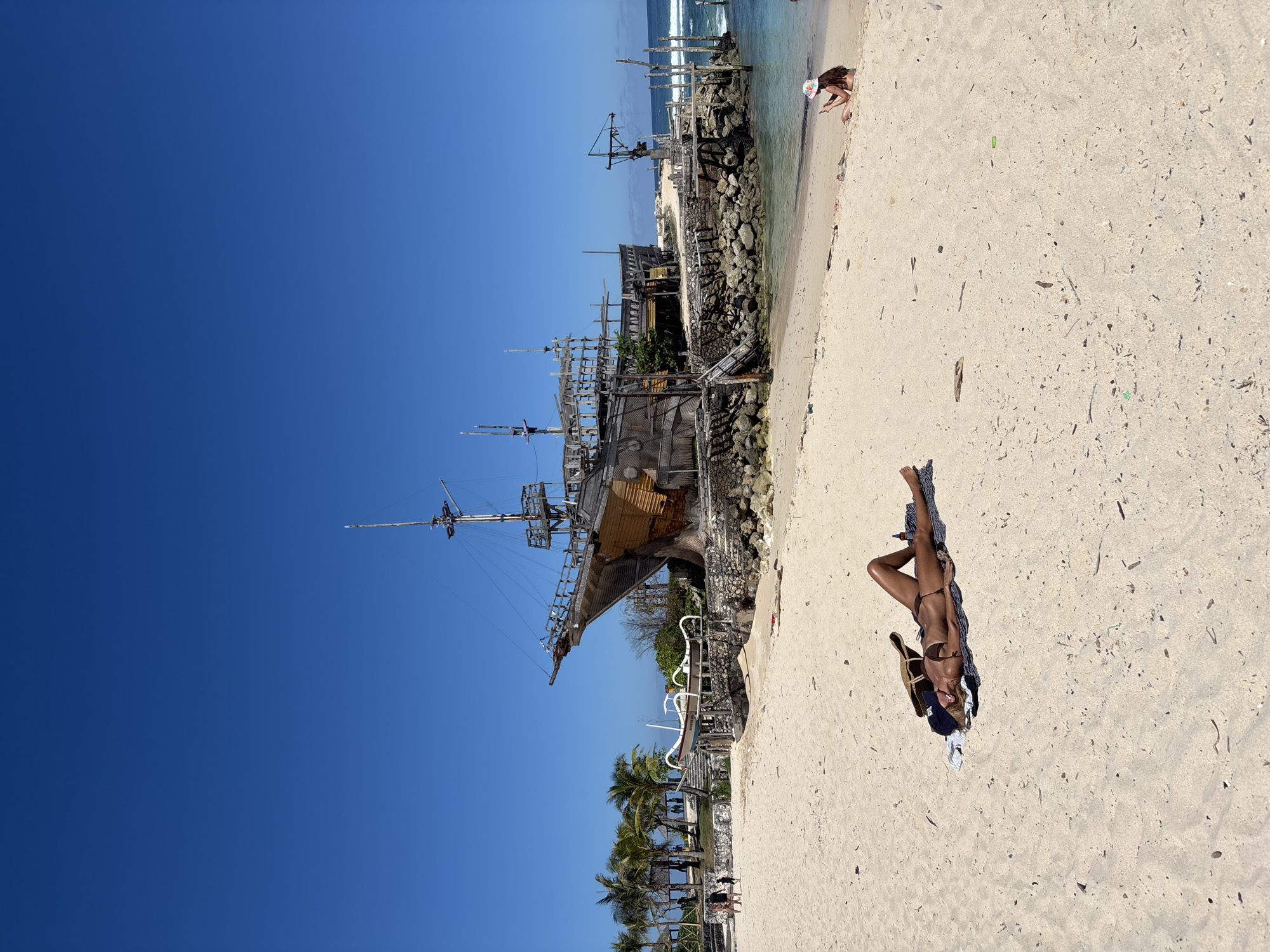 A sunny beach scene in Kuta Selatan, Indonesia. A woman in a brown bikini is lying on a patterned towel on the sand. A partially built pirate ship structure stands nearby. The blue sky is clear. Palm trees and other beachside structures are visible in the background. Some people are on the beach playing in the water.