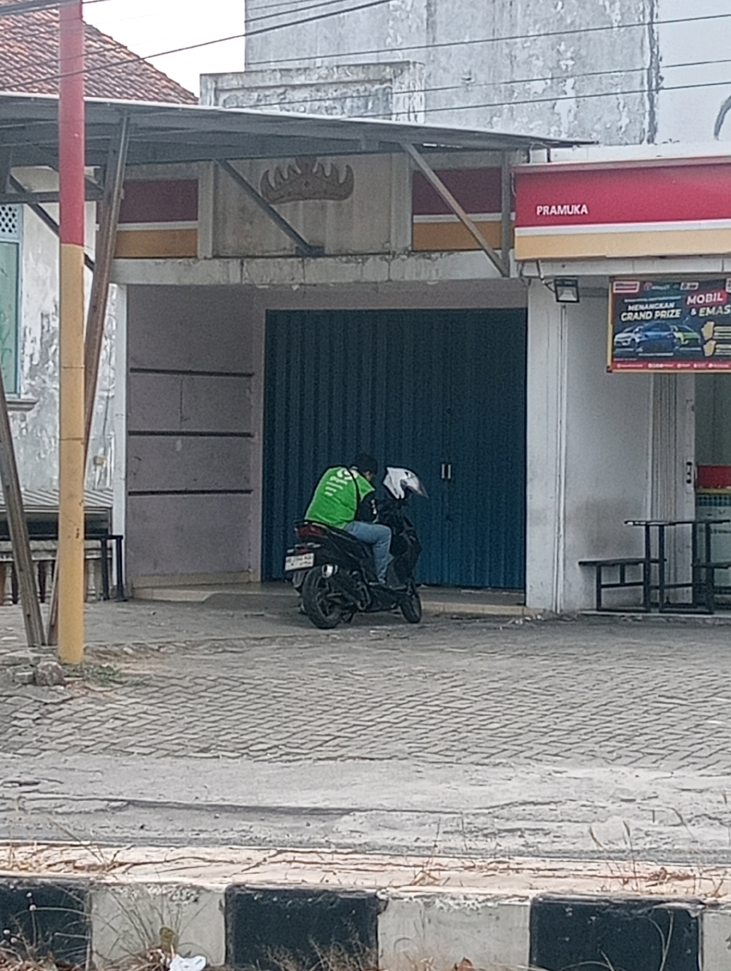 In the city of Bandar Lampung, Indonesia, a person on a black motorcycle with a bright green jacket and backpack is parked on a paved street in front of a building with a large blue roll-up door. A white helmet is on the back of the motorcycle. To the right of the roll-up door, a red and yellow sign reads "PRAMUKA." To the right of the entrance, two black metal tables and benches are set up. The building appears to be a commercial establishment.