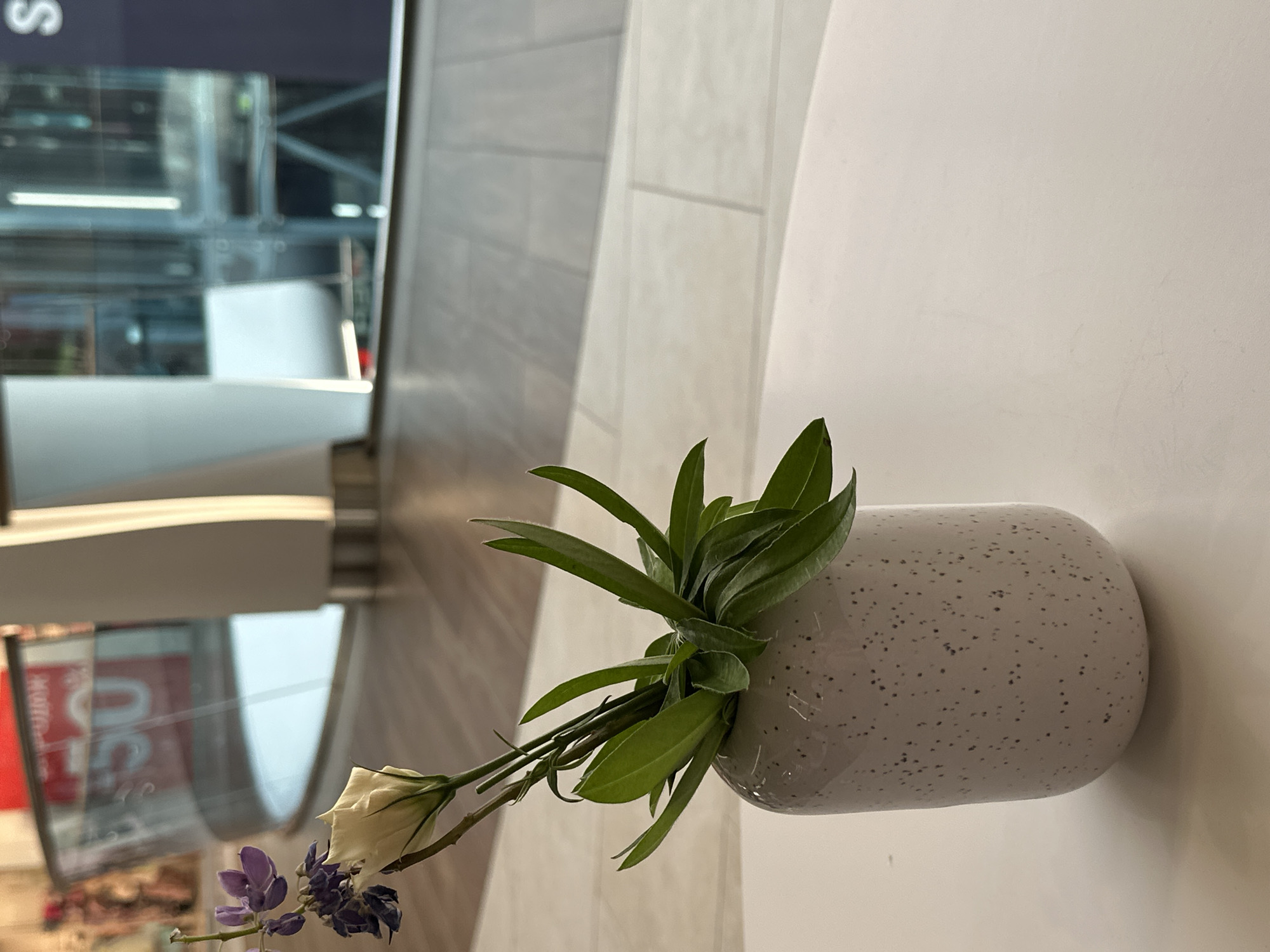 A close-up, eye-level shot focuses on a small, white ceramic vase with dark speckles, resting on a white surface, likely a table. The vase contains a small arrangement of greenery and two flowers: a single, pale cream-colored rosebud and a cluster of small, deep purple flowers. The background suggests an indoor environment, possibly a cafe or lounge area within a public building. The floor is tiled with a wood-grain pattern. In the blurred background, a curved glass railing is visible with a sign displaying "50%" and further on, a dark blue sign with a white letter "S." The lighting is soft, and the overall impression is one of quiet elegance. The scene is devoid of any people, or indications of human activity, and is not specific to Kyiv, Ukraine, or any identifiable city.