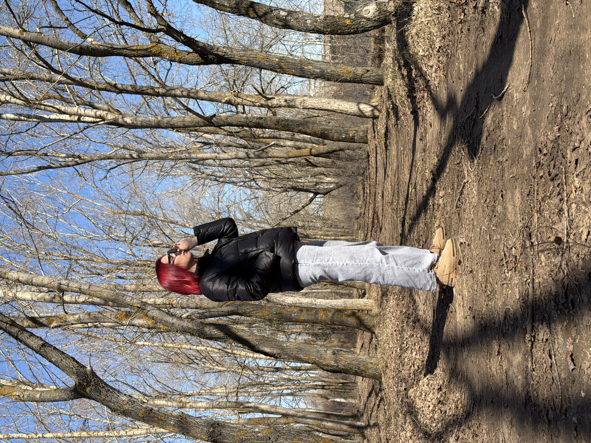 A woman with dark glasses and burgundy hair, wearing a black puffer jacket and grey jeans, stands on a path in a park in Chaikovsky, Russia. The path is lined with trees with bare branches, creating a corridor effect. The ground is covered with dry leaves and soil. The sky is visible through the branches and is blue. The woman is looking upwards and appears to be adjusting her glasses.
