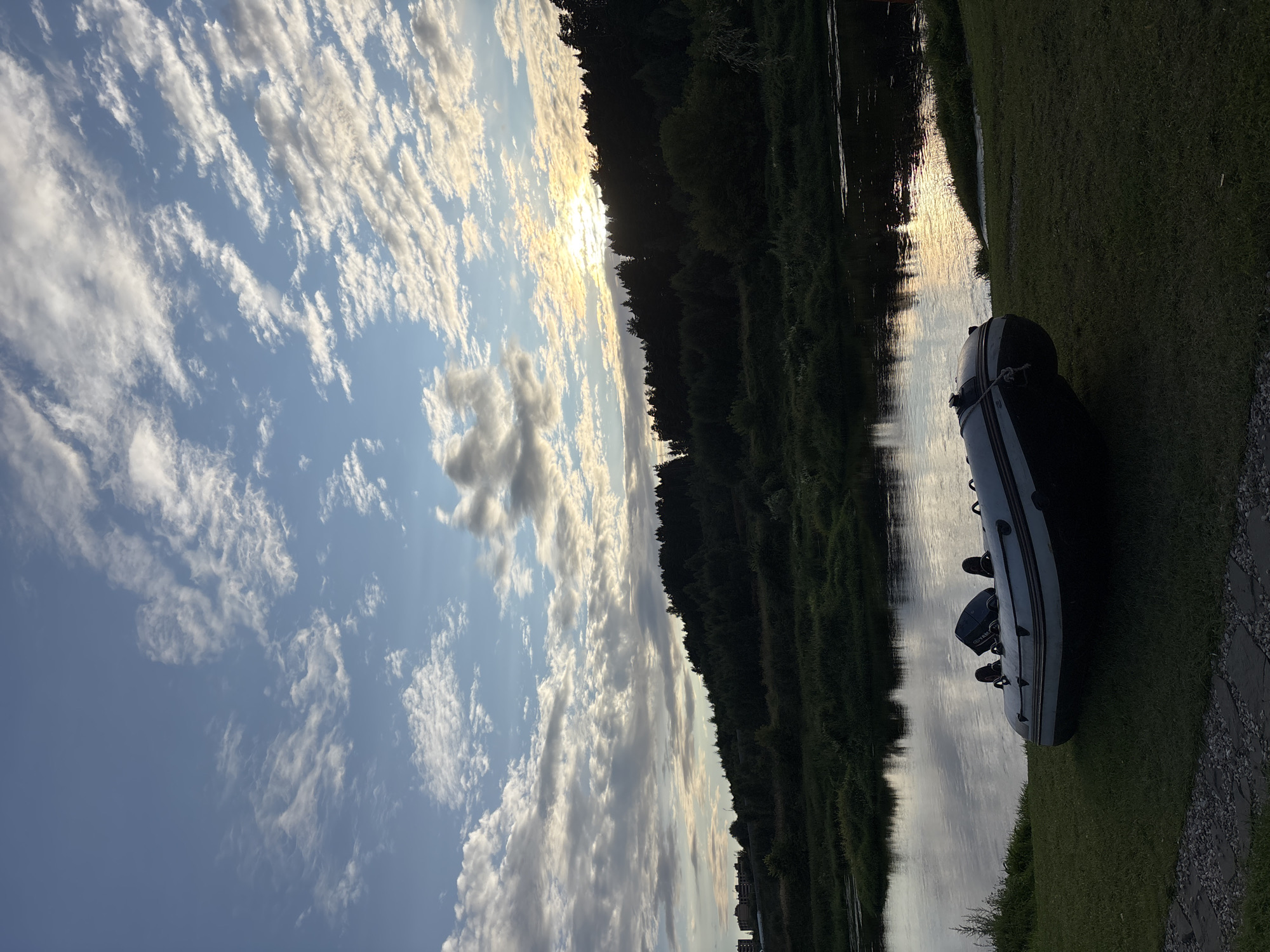 The image shows a calm river scene with an inflatable boat resting on the grassy bank in the foreground. The river reflects the sky, which is filled with scattered clouds and illuminated by the setting sun peeking through the trees on the distant hilly horizon. The far bank of the river is lined with a dense forest. In the far distance on the left side of the image, a cluster of residential buildings is visible. The scene appears to be set in Zvenigorod, Russia, possibly on the Moskva River.
