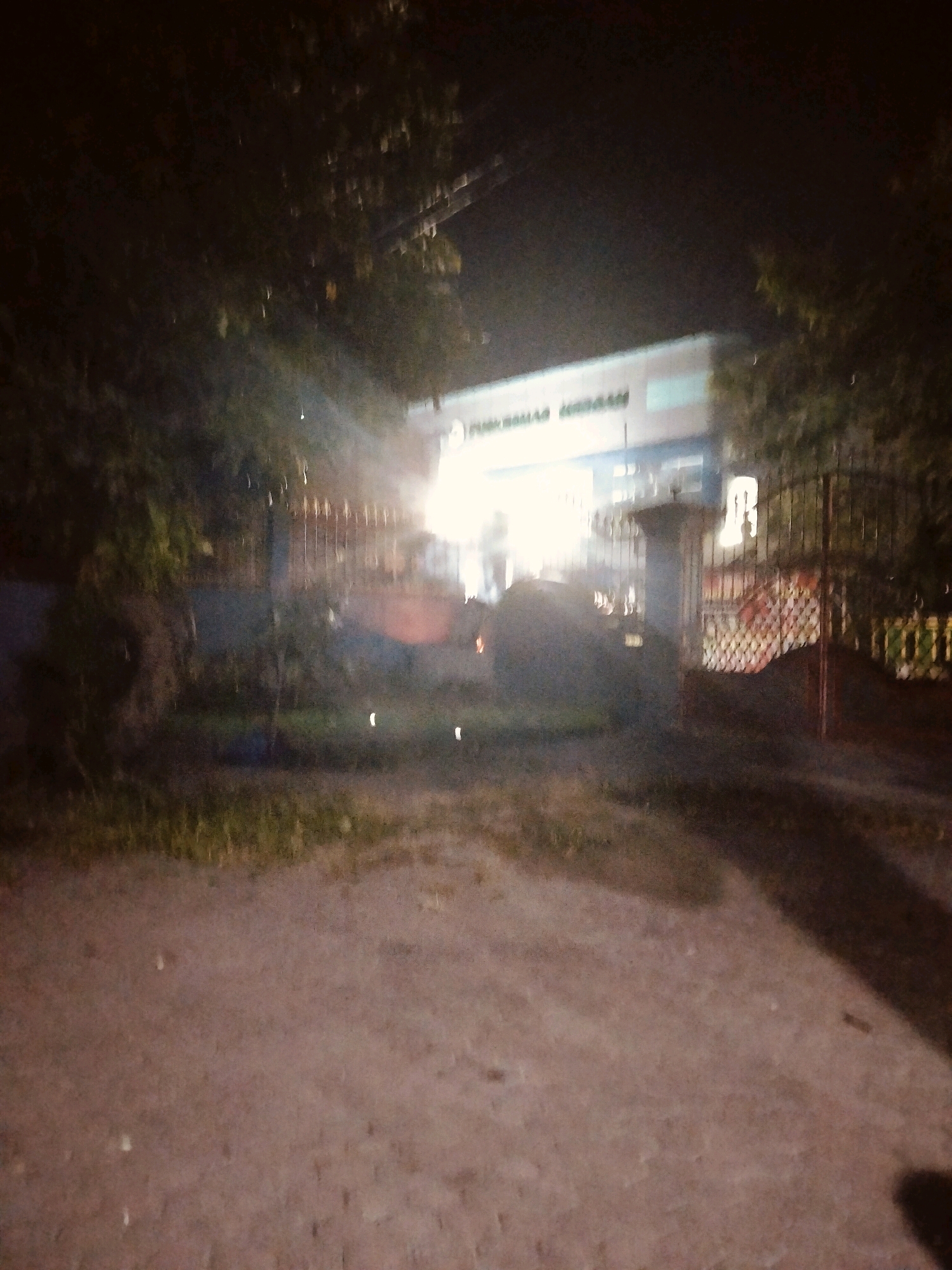 The image depicts the exterior of a building at night, likely in the city of Keboan, Indonesia. A bright light emanates from within or in front of the building, illuminating a portion of the facade and the surrounding area. The building appears to have signage with text that is partially legible, suggesting it could be a public institution or business. A metal fence runs across the front of the property, with a gate visible. Trees and foliage are present on either side of the building and in the foreground. The ground is a mixture of dirt and sparse grass. The overall impression is of a dimly lit, possibly active, location after dark.