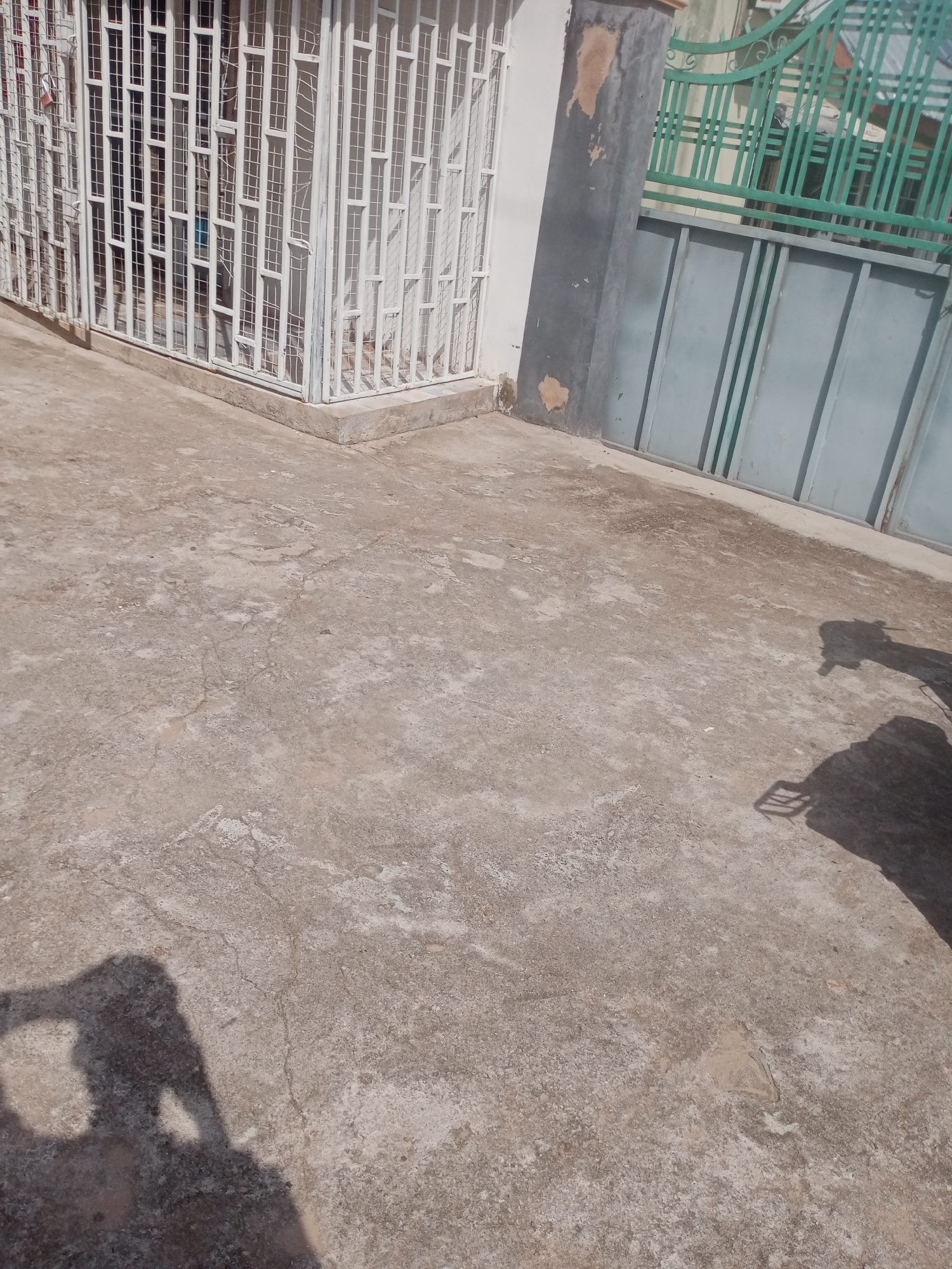 The image shows a concrete ground in front of a building with a white metal grate and a green metal gate. There are shadows of two people on the ground, suggesting they are standing nearby, possibly taking the photo. The scene appears to be an outdoor space in the city of Jalingo, Nigeria, possibly a residential or commercial area. The concrete ground is uneven and shows signs of wear and tear, with cracks and patches of discoloration. The white metal grate covers windows or openings on the building, and the green metal gate appears to be part of a fence or entrance. The overall impression is of a functional, everyday outdoor environment.