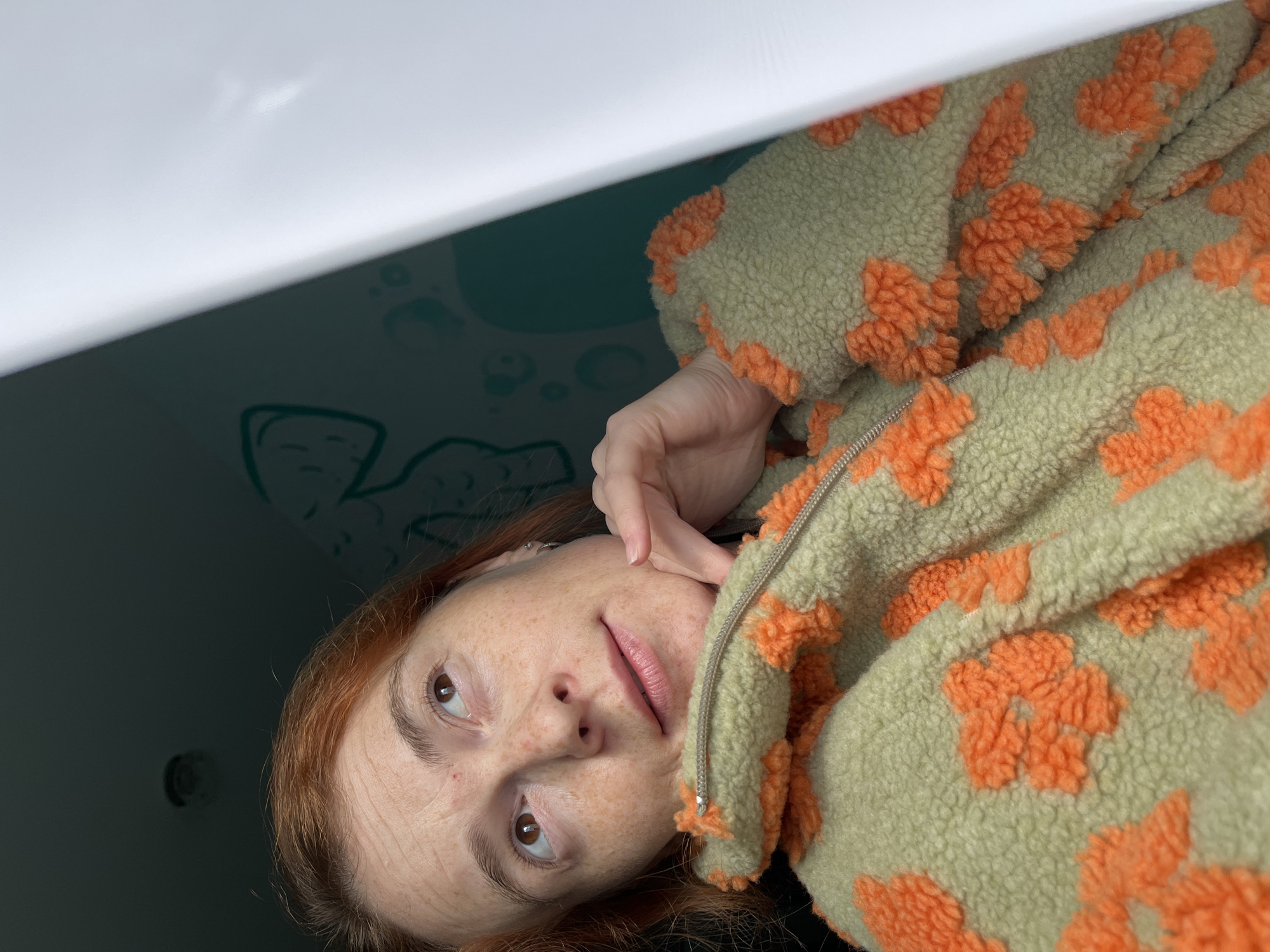 A medium shot captures a pale-skinned woman with auburn hair and freckles, looking upwards. She is wearing a light green fuzzy jacket with a bold orange floral pattern. Her right hand is gently resting under her chin, and her left arm is obscured by the jacket. The background shows a teal wall with faint green outlines of abstract shapes and a dark ceiling with a light fixture. A bright white surface, possibly a wall or window frame, is visible on the right side of the frame, partially obscuring the background. The image appears to be taken indoors, possibly in a domestic setting, and there is no indication of the city of Chaikovsky, Russia.