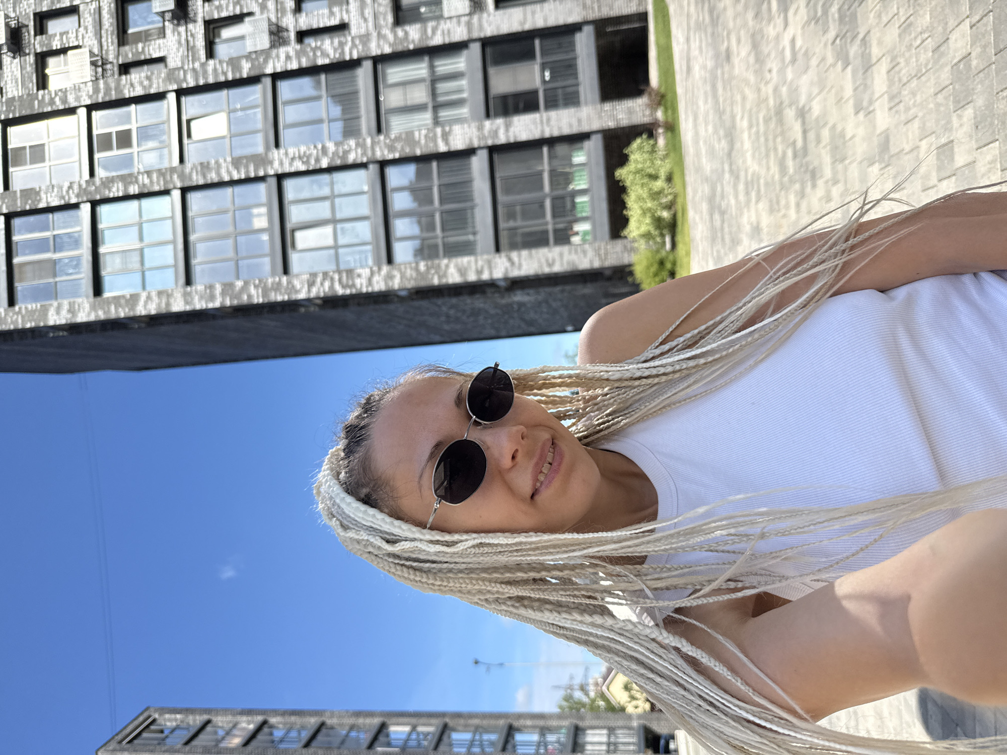 In this waist-up, selfie-style outdoor photograph, a woman with blonde braided hair and dark oval sunglasses is positioned in the foreground, looking towards the camera with a slight smile. She is wearing a white tank top. The background features two modern, dark-colored apartment buildings. The building to the right is larger and closer to the camera, showcasing numerous windows and balconies. The building to the left is further away and appears similar in architectural style. The environment is an urban setting with a paved walkway and some greenery visible in the mid-ground. The sky is clear and blue, indicating a sunny day. The image appears to have been taken in the city of Rusanovo, Russia.