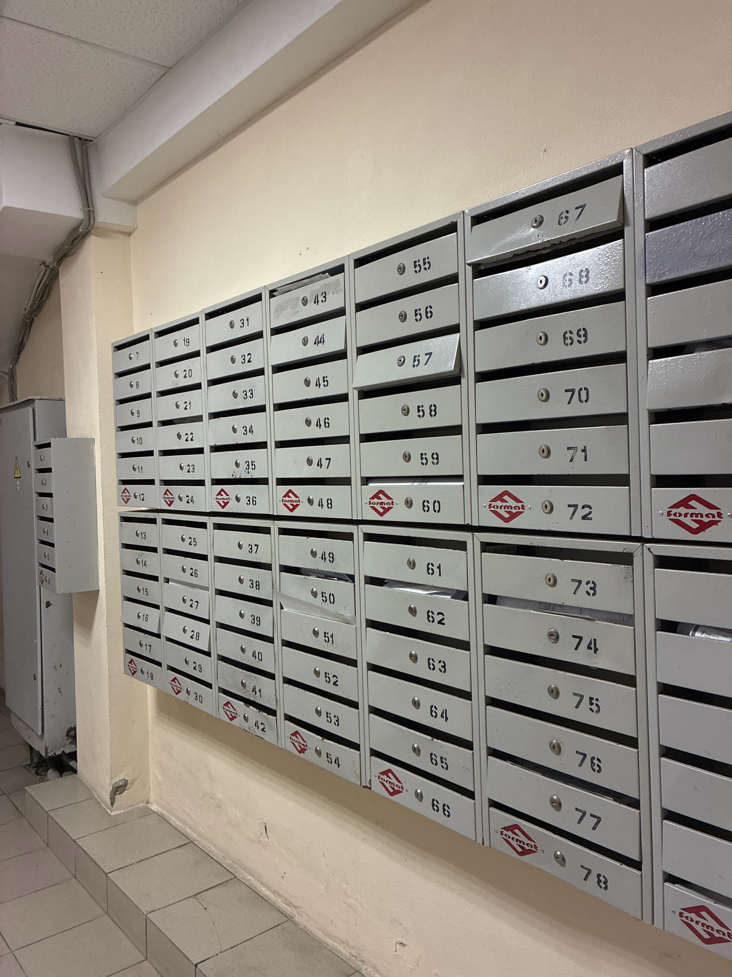 The image shows a wall of metal mailboxes in an apartment building hallway. The mailboxes are arranged in vertical columns and horizontal rows. Each mailbox is numbered, with numbers ranging from 7 to 78 visible. Some of the mailboxes appear to be open, with mail visible inside. There is a large grey electrical box on the left side of the image, attached to the wall. The walls are painted a light cream color, and the floor is tiled with light grey tiles. The lighting is fluorescent and appears to be from overhead fixtures. The scene is clean and well-maintained, suggesting it is a typical hallway in a residential building. It is not possible to determine the exact location within Sochi, Russia, from the image alone, but the style of the mailboxes and building is common in many Russian apartment complexes.