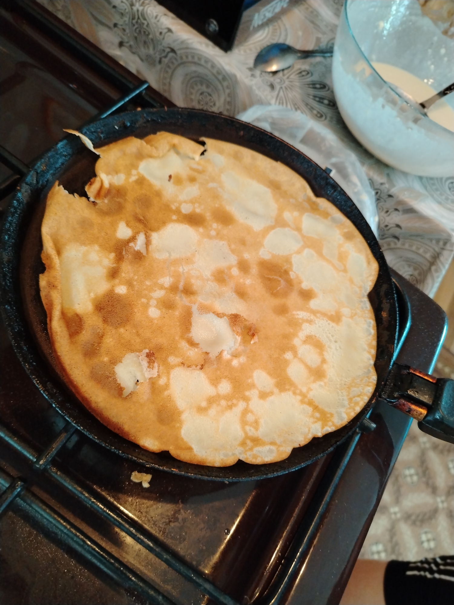 A pancake is being cooked in a black frying pan on a gas stovetop. The pancake is mostly golden brown with patches of lighter color. Next to the stove is a glass bowl containing pancake batter. A spoon is visible in the bowl. A patterned surface is visible in the background. The location is possibly Большая Уса, Russia.