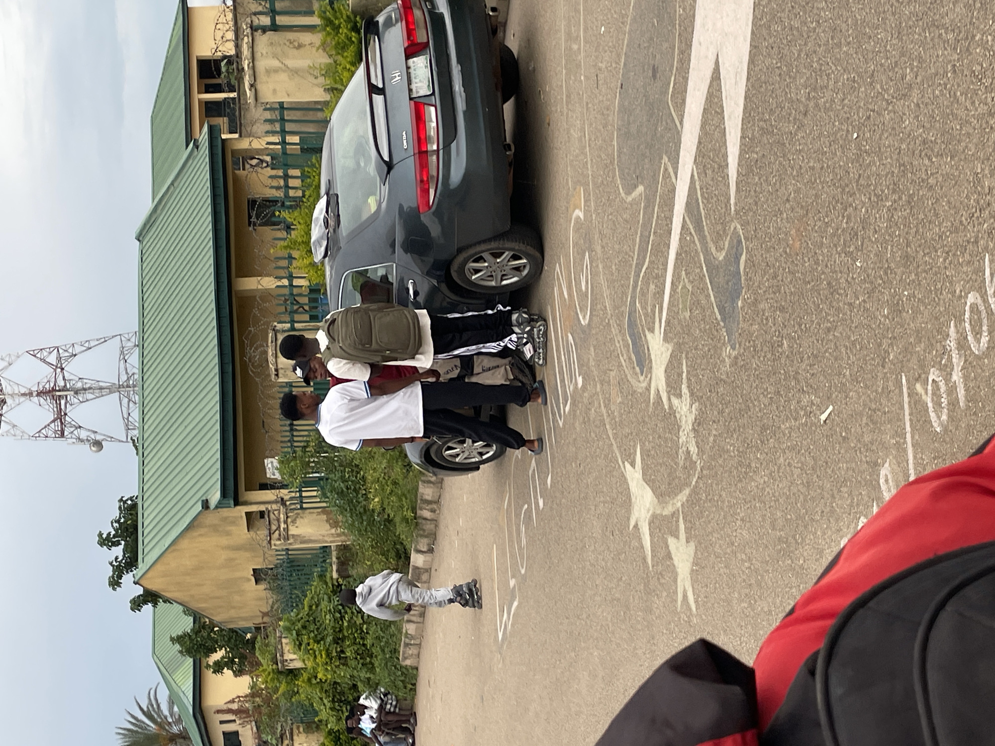 In Jos, Nigeria, a group of young men are gathered near a dark-colored sedan parked on an asphalt road. One individual is rollerblading, facing away from the camera. Two others, dressed in casual attire, stand near the car. The background features residential buildings with green roofs, a fence, and lush vegetation. A tall communication tower is visible in the distance, with a cloudy sky overhead. The road surface has chalk drawings of figures and stars.