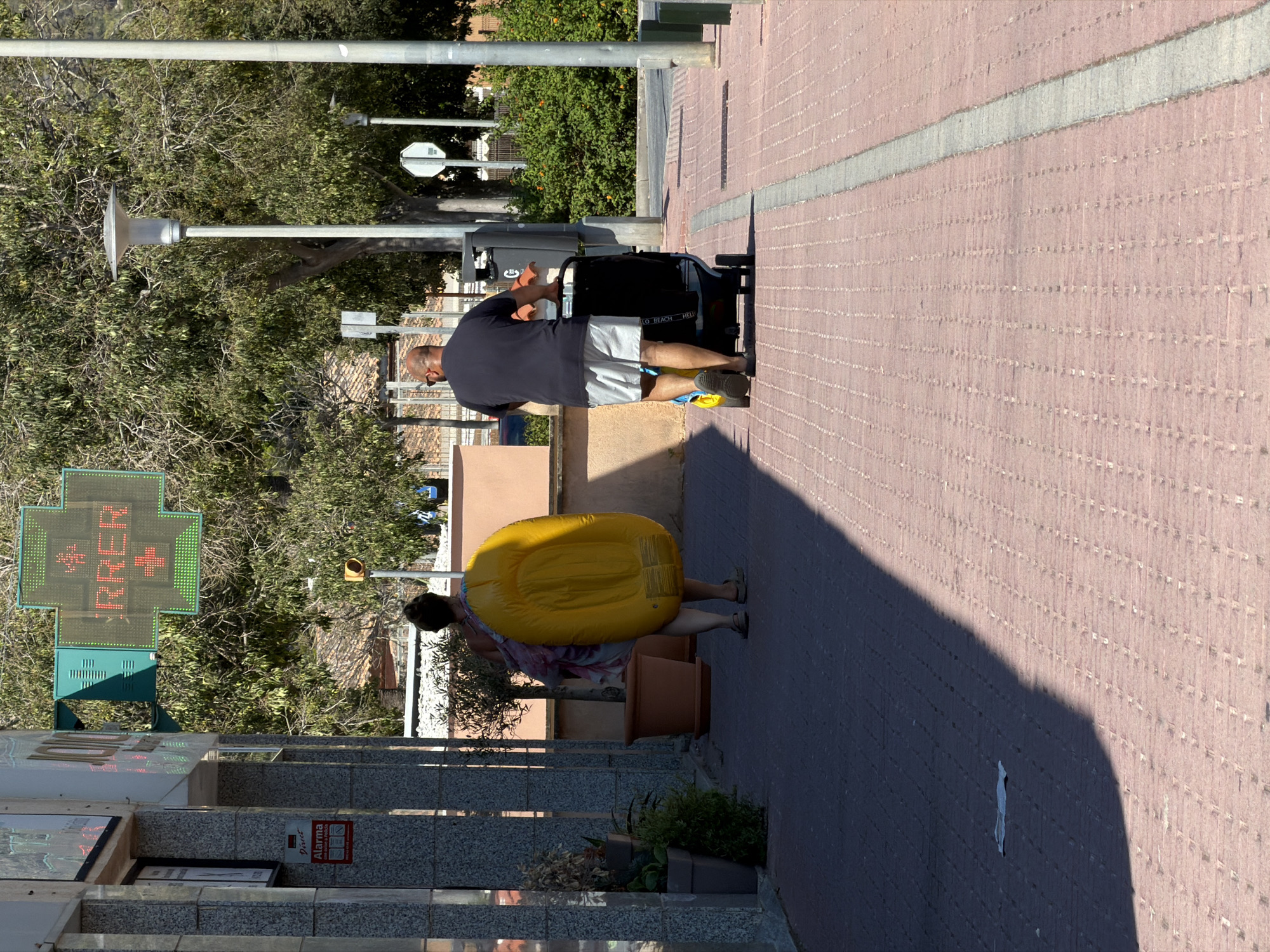 The image shows a street scene in Calvià, Spain, on a sunny day. In the foreground, a man with a shaved head is pushing a dark-colored stroller with a red accent along a paved sidewalk made of red brick pavers with a light gray dividing line. He is wearing a dark blue t-shirt and light gray shorts. A woman is walking in front of him, carrying a large, deflated, bright yellow inflatable raft on her right side. She is wearing a patterned dress or wrap and dark sandals. To the left, a building with a gray stone facade has a red alarm sign visible. Above the building, a green pharmacy cross sign with red LED lights displays the word "RRER". In the background, there are trees with green foliage, a street lamp, and other street elements, suggesting an outdoor commercial or residential area. The overall impression is a candid shot of people going about their day in a Mediterranean setting.
