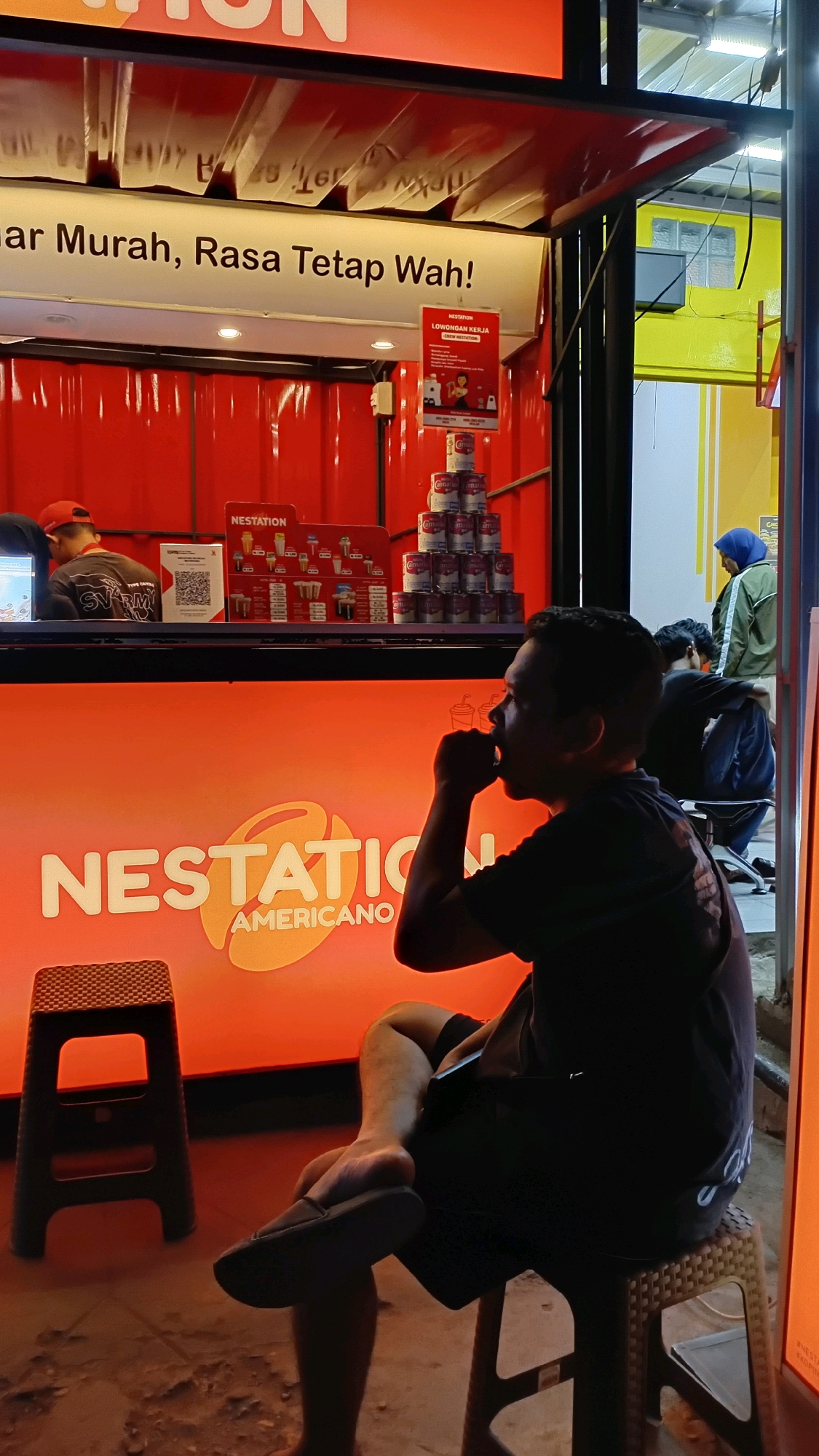 The image depicts a nighttime scene outside a food stall, likely in the city of Jambi, Indonesia. The stall features a bright orange counter with "NESTATION AMERICANO" prominently displayed. Behind the counter, a menu board shows various drink options, and a stack of cans is visible. A "LOWONGAN KERJA" (Job Vacancy) sign is also posted. In the foreground, a man in silhouette is seated on a plastic stool, drinking from a cup. He is wearing a dark t-shirt and shorts, with his right leg crossed over his left. To his left, another plastic stool stands empty. The background shows a yellow building with some windows and a glimpse of a person wearing a blue hijab and green jacket. The overall atmosphere suggests a casual outdoor dining or takeaway establishment operating after dark.